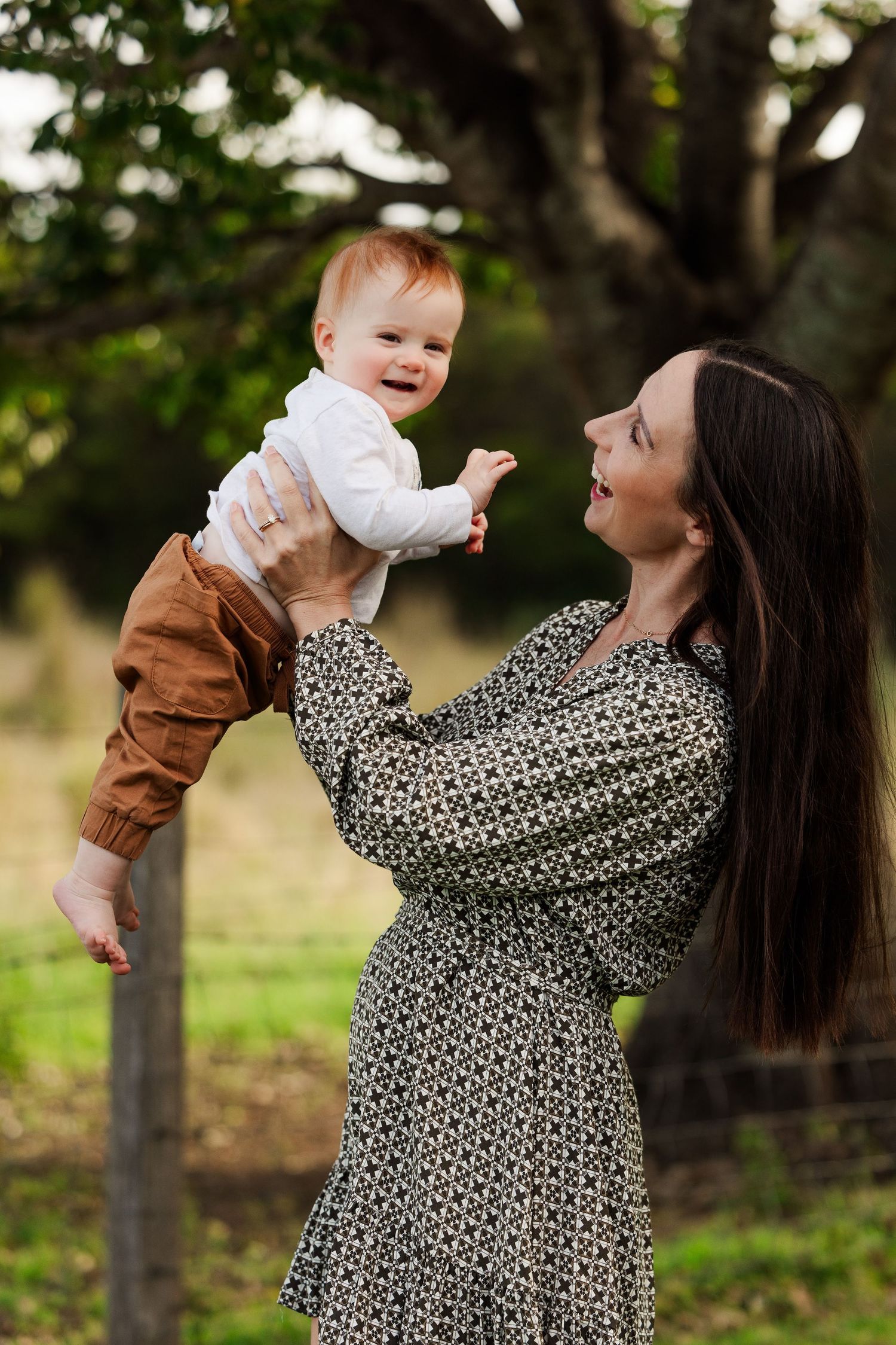 A joyful mother lifts her smiling red-haired baby outdoors, surrounded by lush green trees.