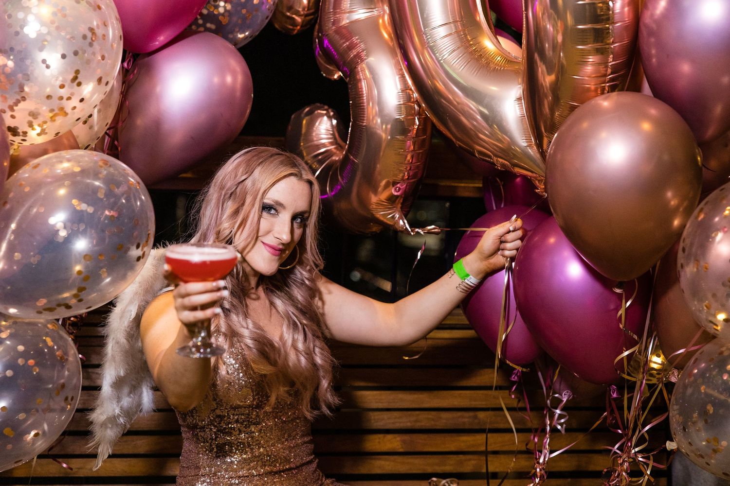 Person in sparkly dress celebrates with cocktail surrounded by pink and gold balloons at festive party.