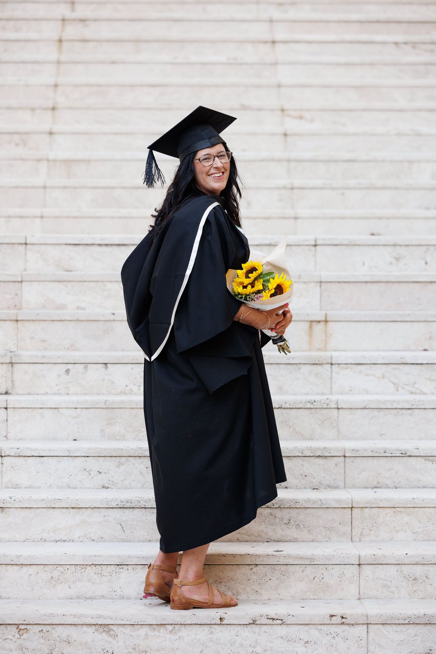 Graduate in black cap and gown holding sunflowers stands joyfully on white stone steps during graduation ceremony.
