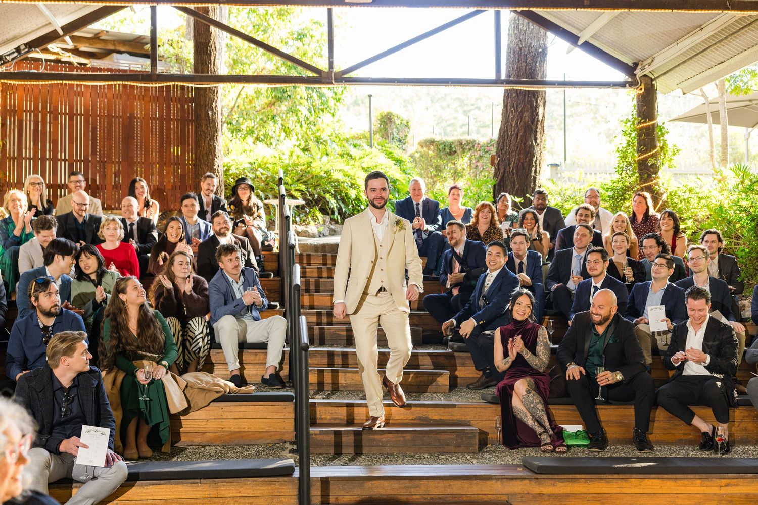 A well-dressed man in a beige suit walks down steps before a seated crowd at an elegant outdoor wedding ceremony.
