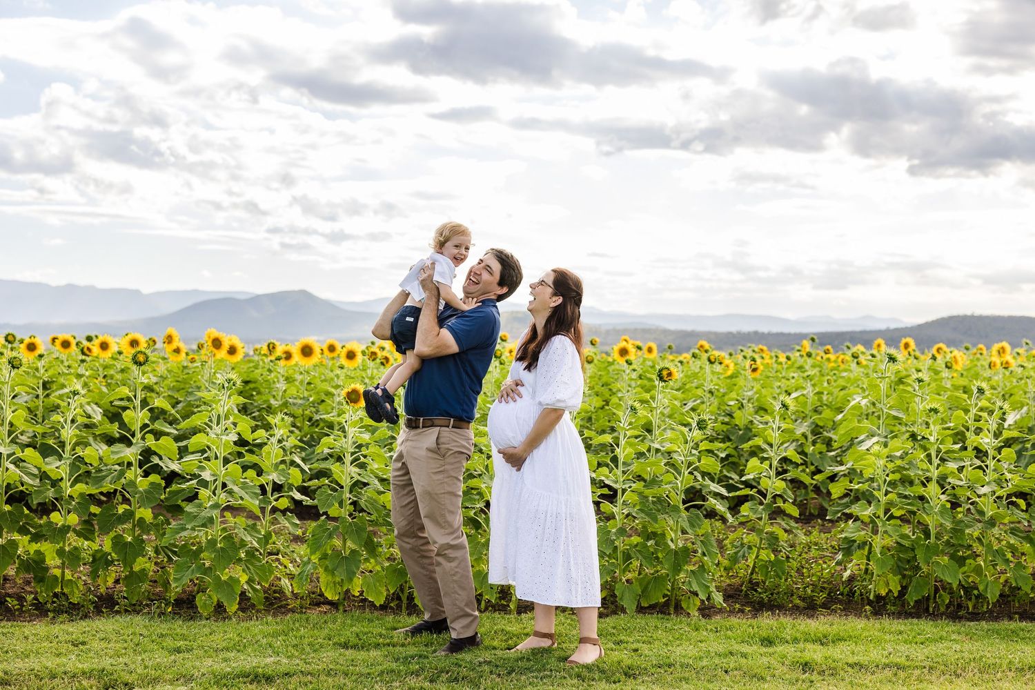 A family shares a tender moment together while standing in front of a vibrant sunflower field under cloudy skies.