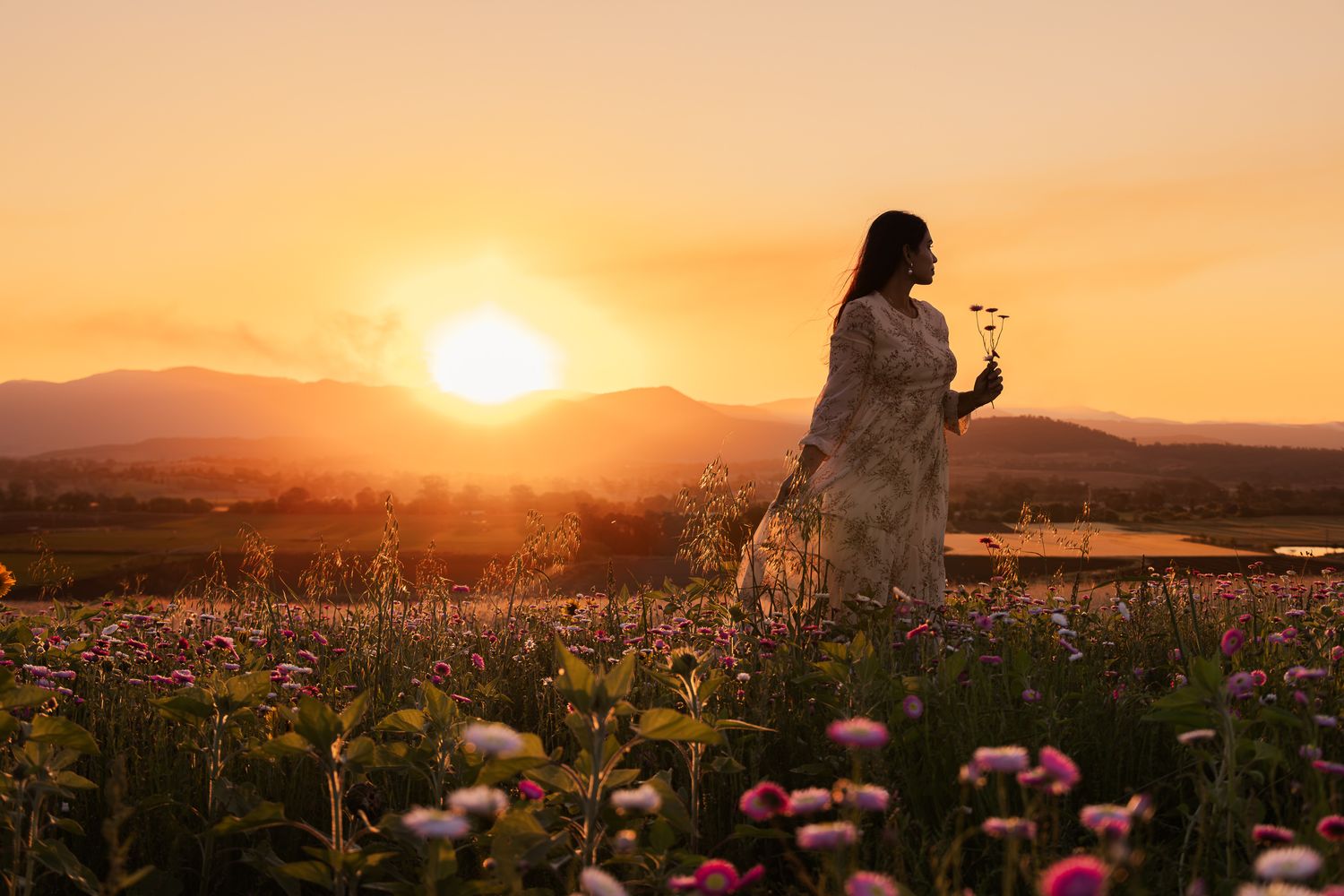 Woman in floral dress standing in a blooming flower field during a golden sunset with mountains in the background.