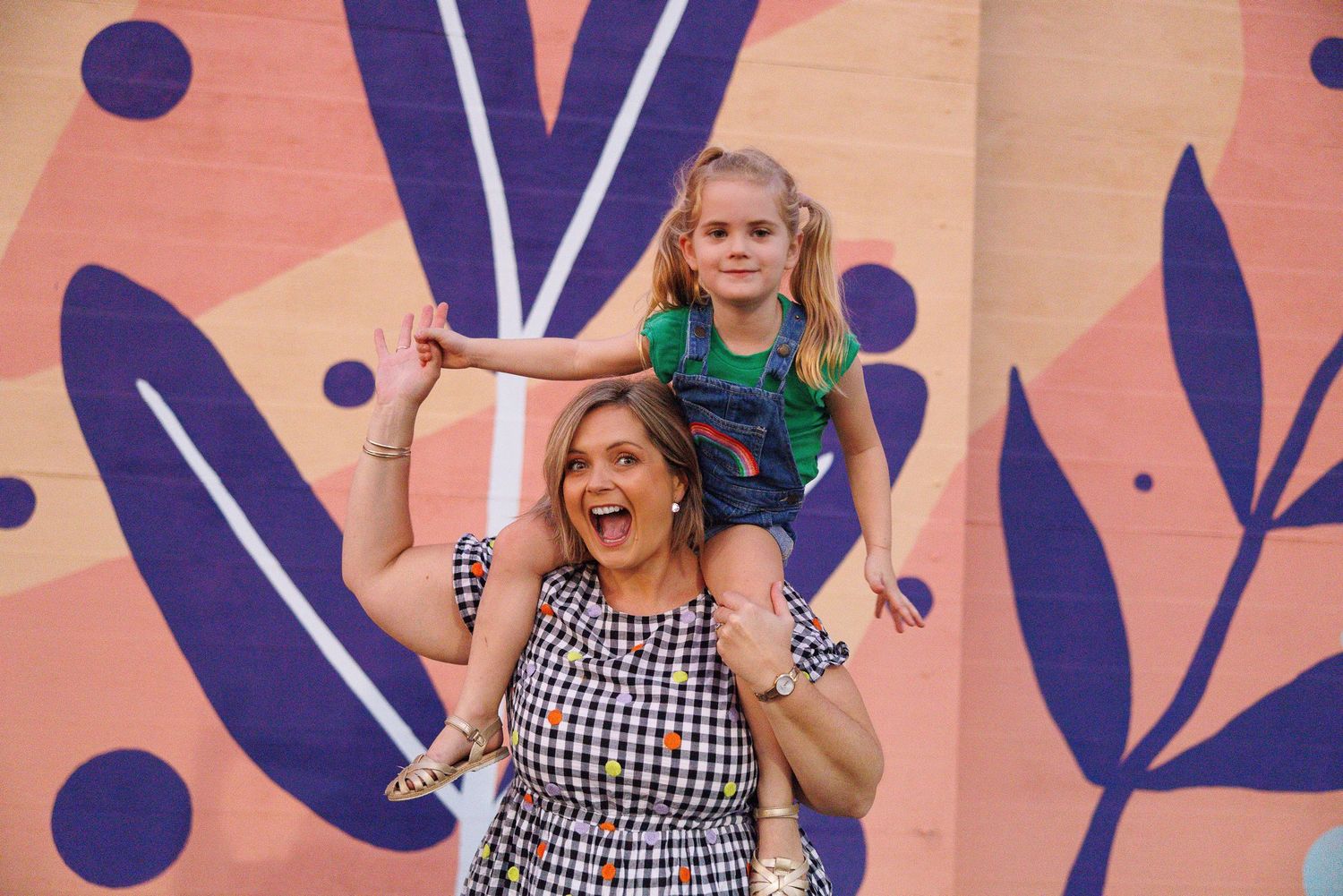 A person in a black and white gingham dress poses expressively against a pink floral background.
