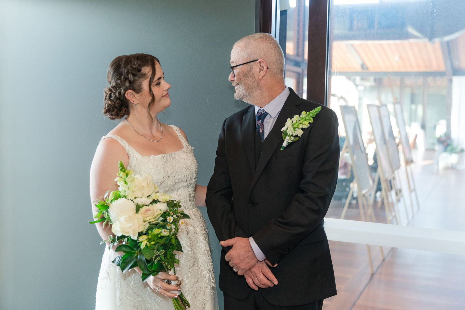 Bride holding white bouquet stands with groom in black suit near window at indoor wedding venue.