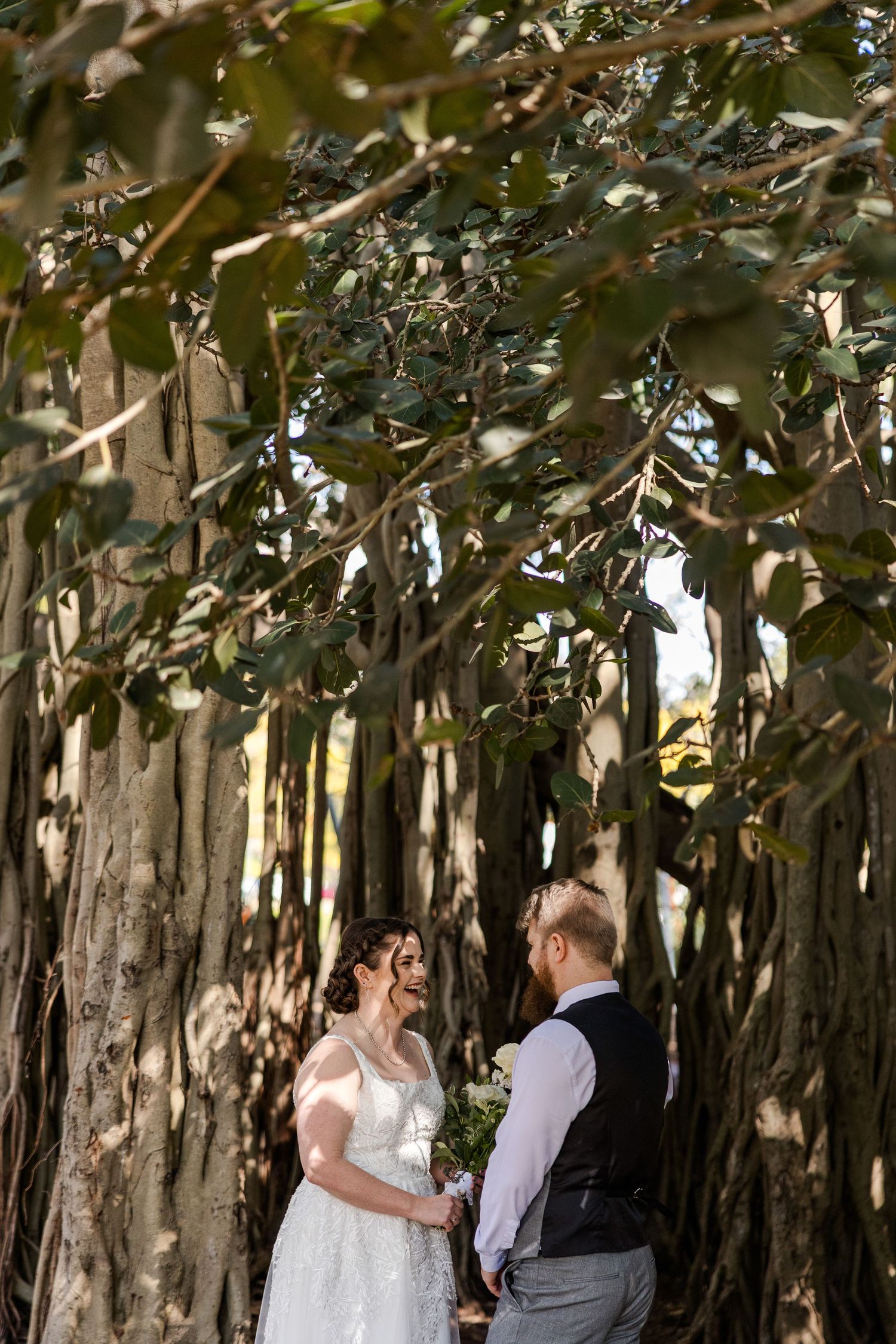Bride and groom hold hands under sprawling banyan tree canopy during outdoor wedding ceremony.