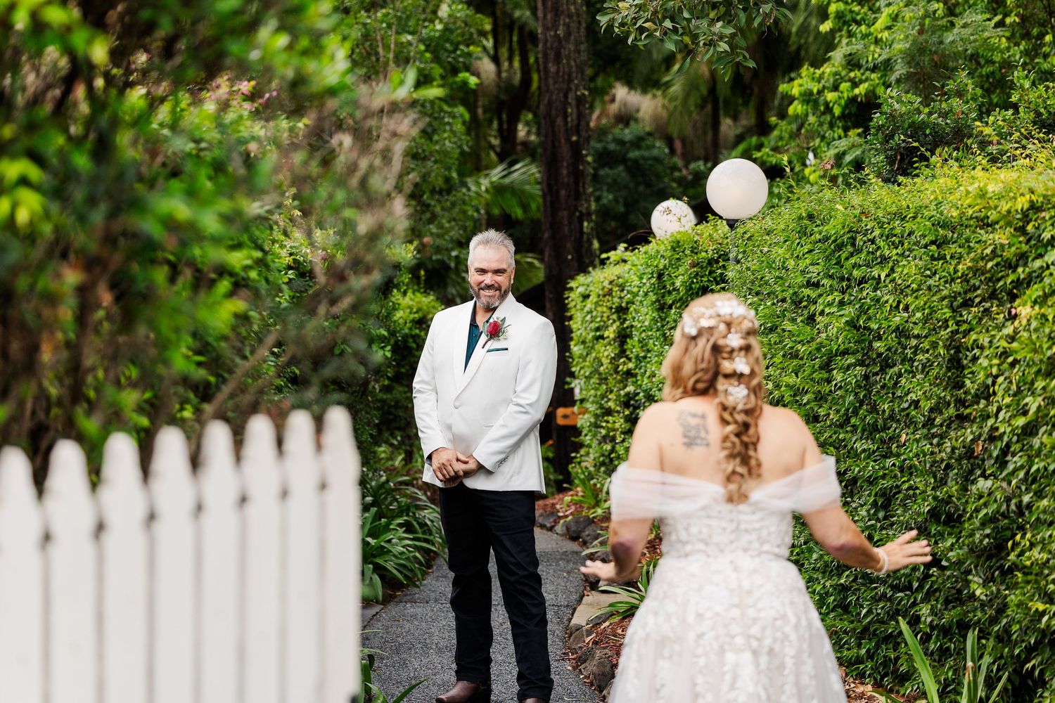 Groom in white suit watches bride approach during first look in a lush garden setting with white picket fence.