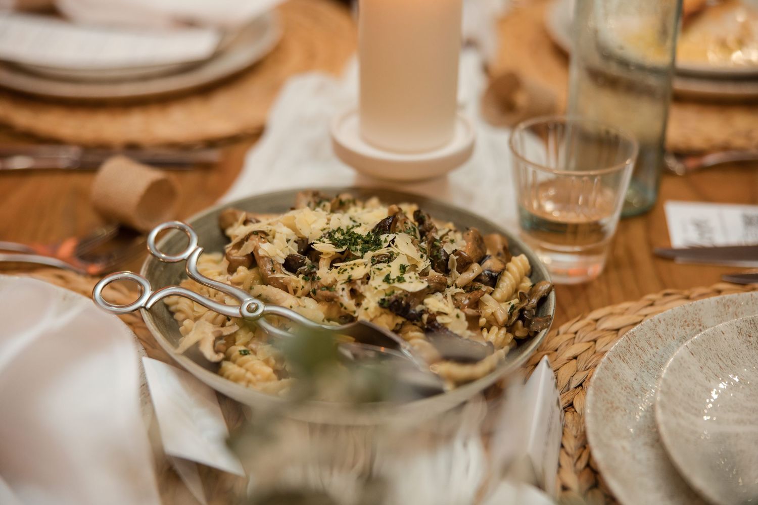 Elegant table setting with a silver serving dish of mushroom risotto garnished with herbs, surrounded by glasses and plates.
