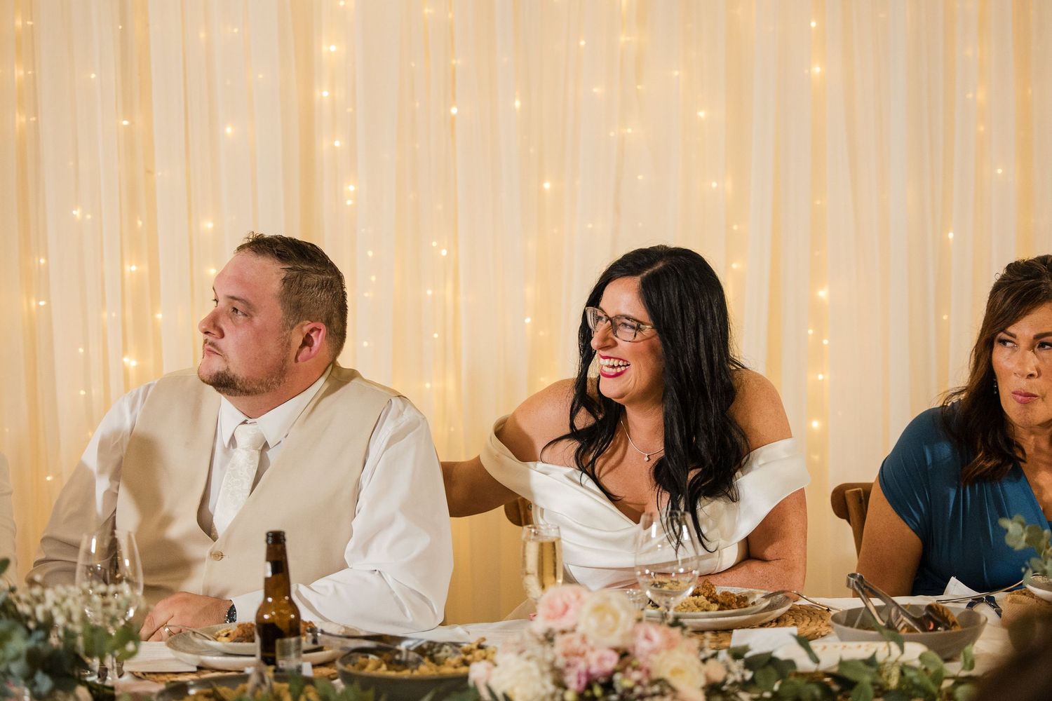 Bride and groom laughing at wedding reception dinner table with fairy light backdrop.