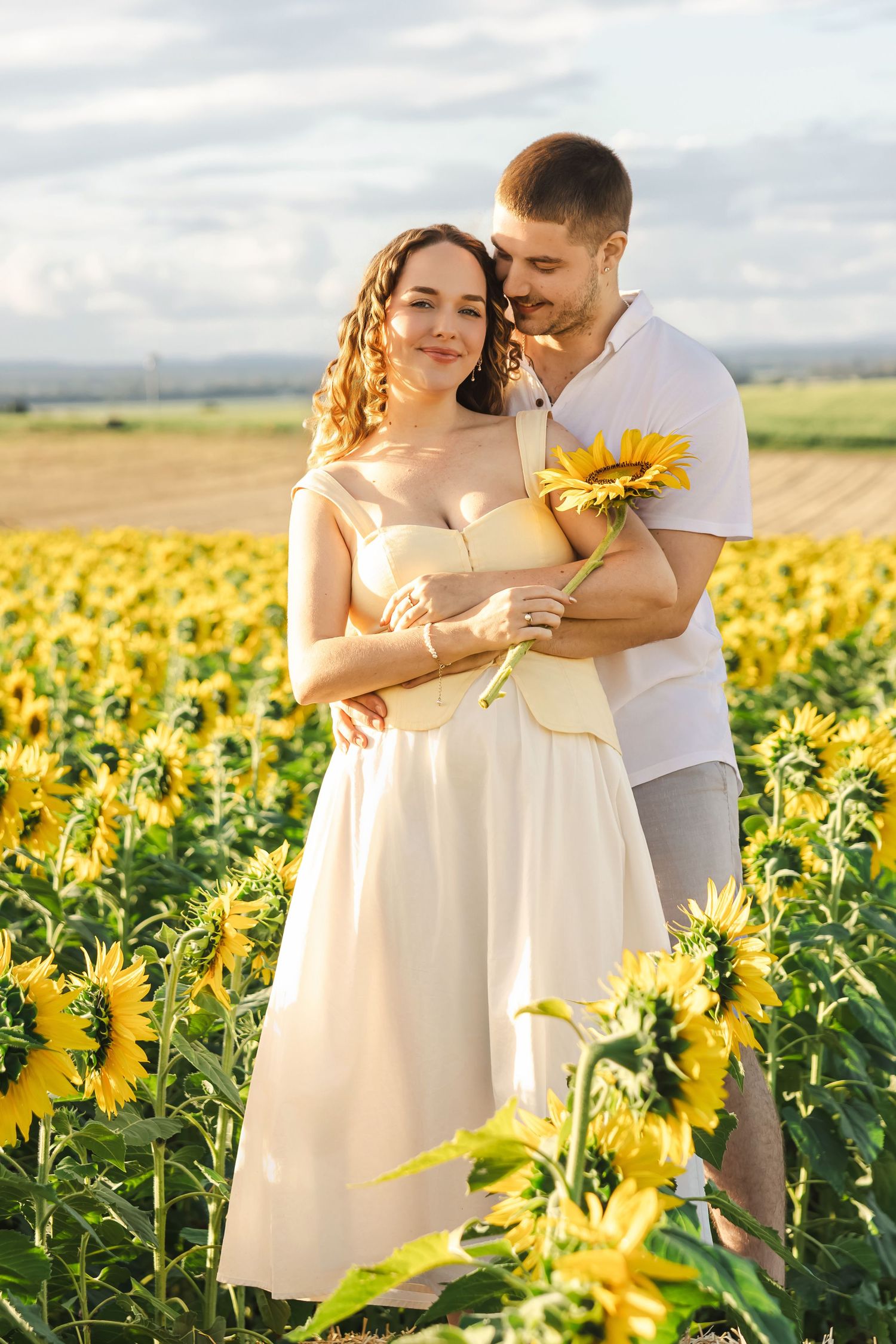 Romantic couple embracing in a sunflower field, woman in white dress holding a sunflower at golden hour.
