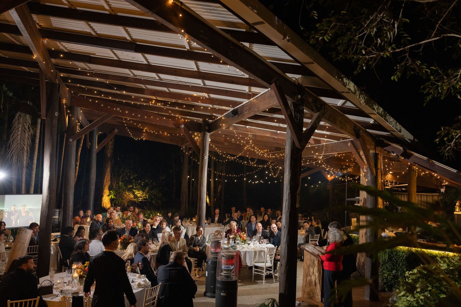 Elegant outdoor wedding reception under a wooden pergola with string lights at night, guests dining at decorated tables.