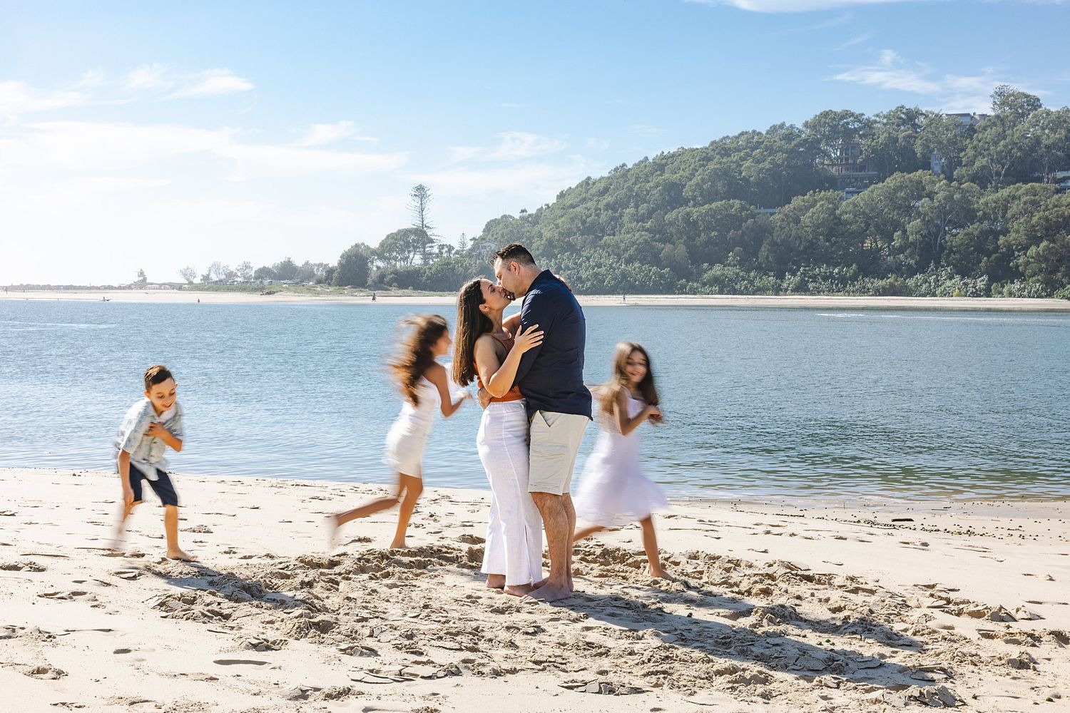 A group enjoys a sunny day playing and walking together on a sandy beach with blue water and hills in the background.