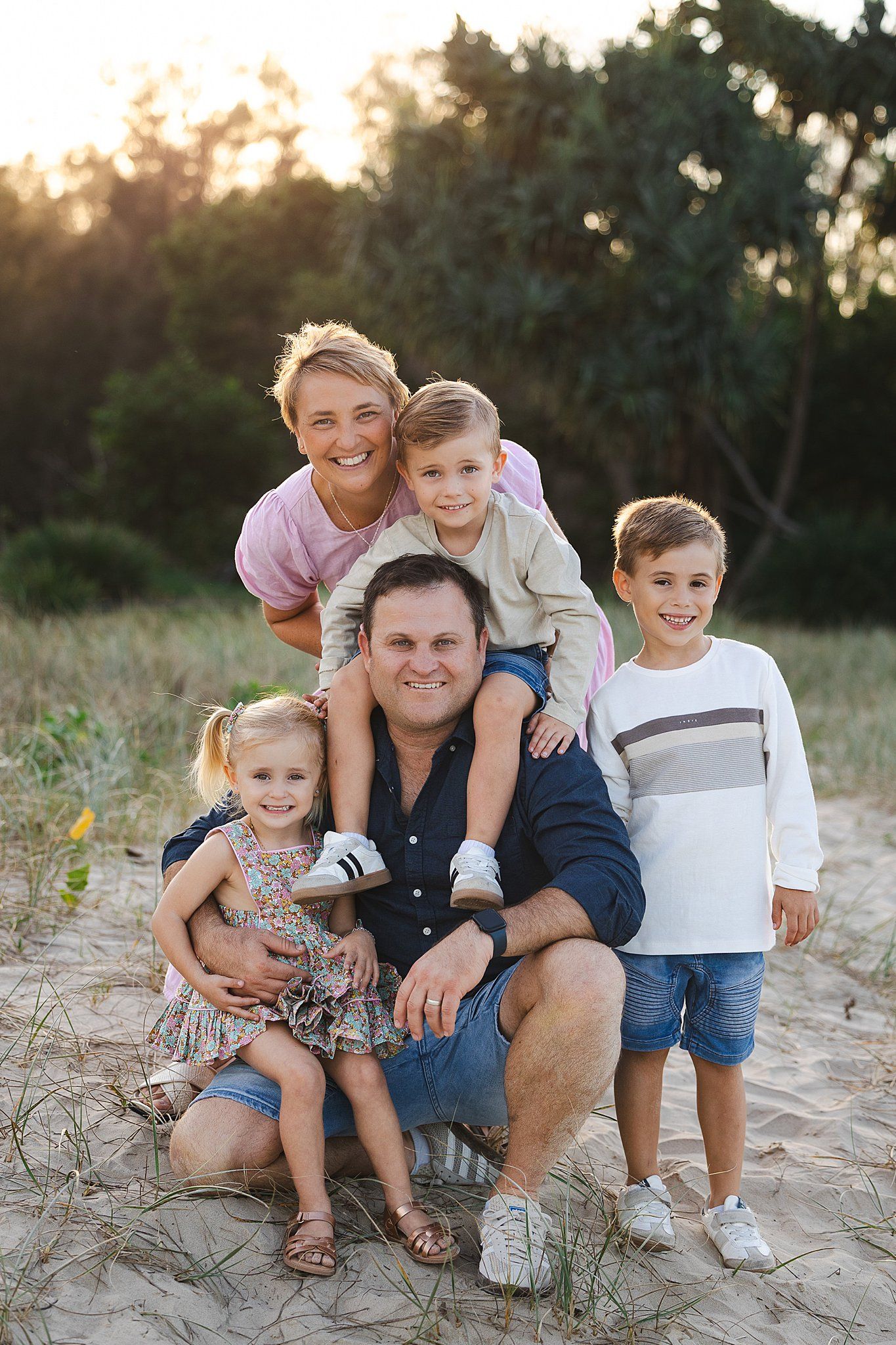 Happy family poses together outdoors on sandy ground at sunset with trees in background.