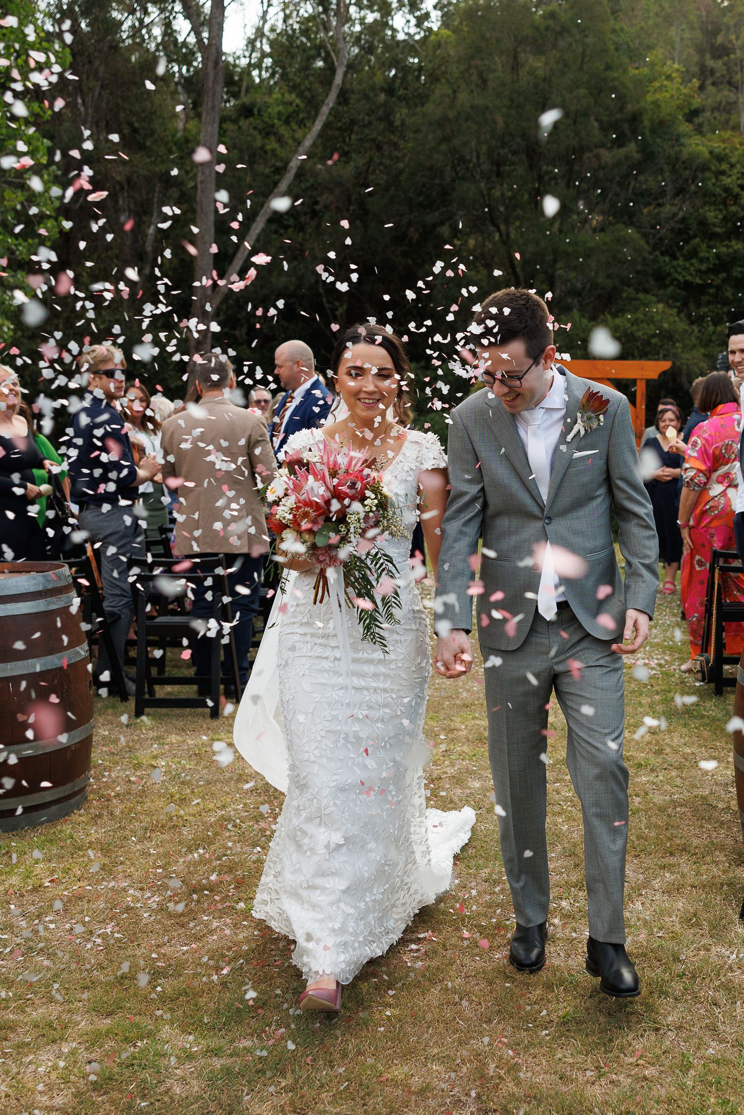 Bride and groom walk through confetti shower after outdoor wedding ceremony, bride holding bouquet.