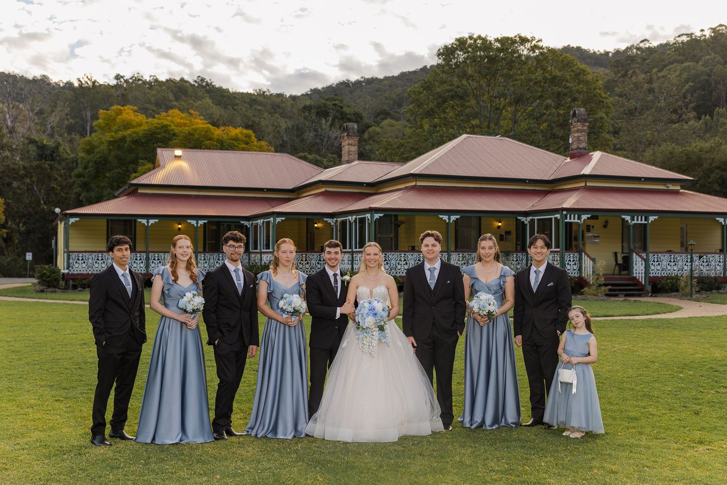 Wedding party in dusty blue attire poses before a historic Queensland homestead with mountain backdrop.