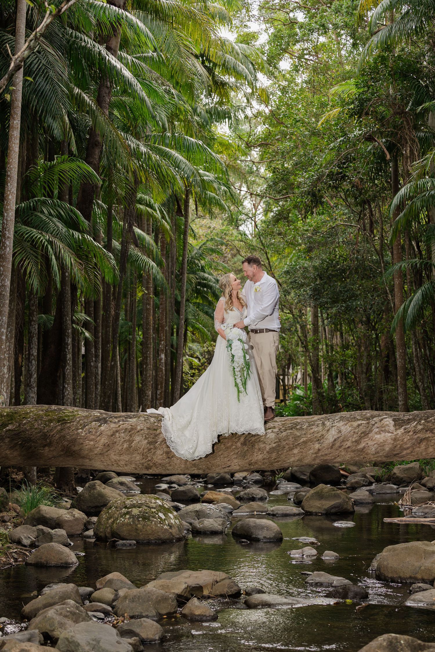 Bride and groom share a romantic moment on a fallen log over a creek surrounded by lush tropical rainforest palms.