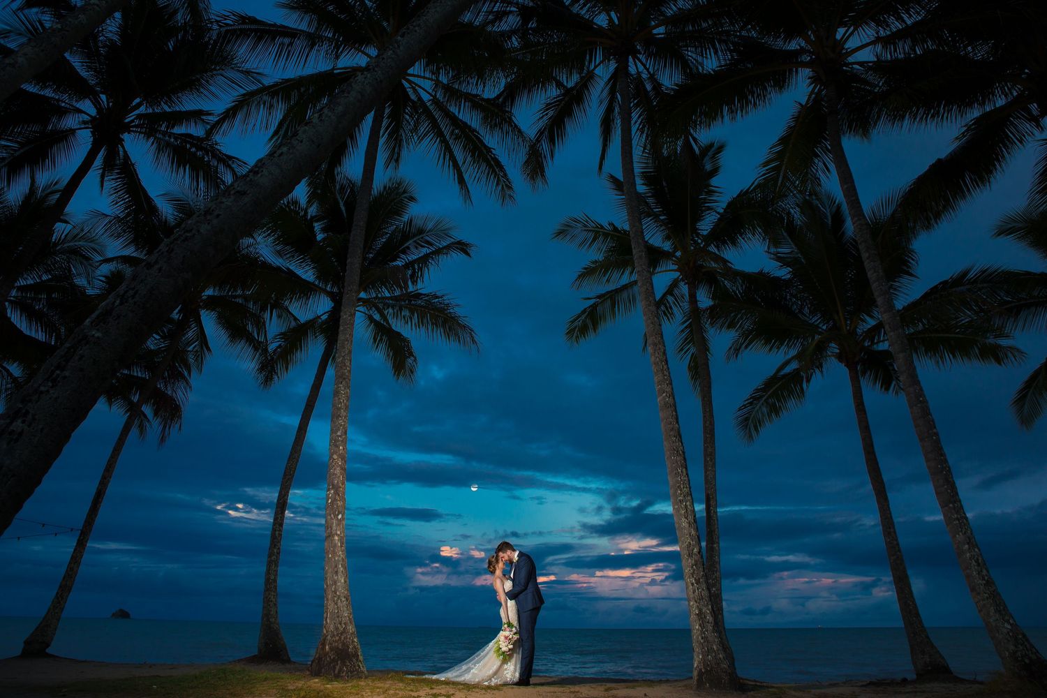 A couple embraces under tall palm trees against a dramatic blue twilight sky at a tropical beach wedding.