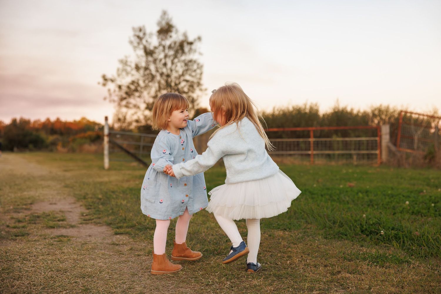 Two young children in light-colored dresses dance together playfully in a grassy field at sunset.