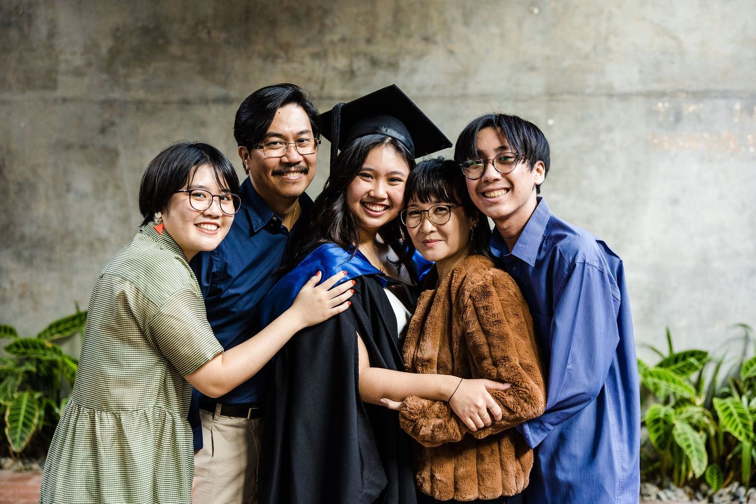 Joyful group hugging and celebrating a graduation ceremony in front of a concrete wall with green plants.