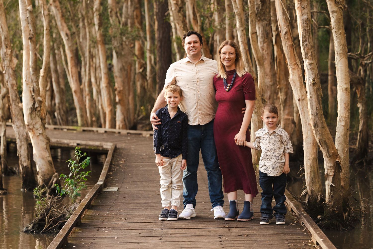 Family group poses on wooden boardwalk path surrounded by tall paperbark trees in natural setting.