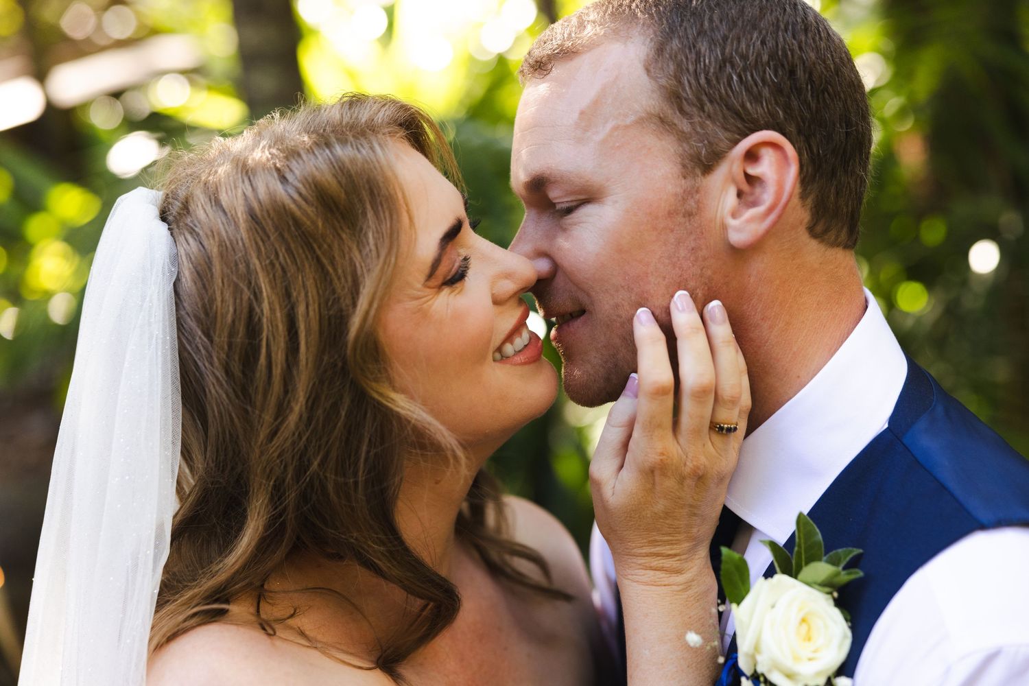 A bride and groom share an intimate moment outdoors with soft bokeh lighting and greenery in the background.
