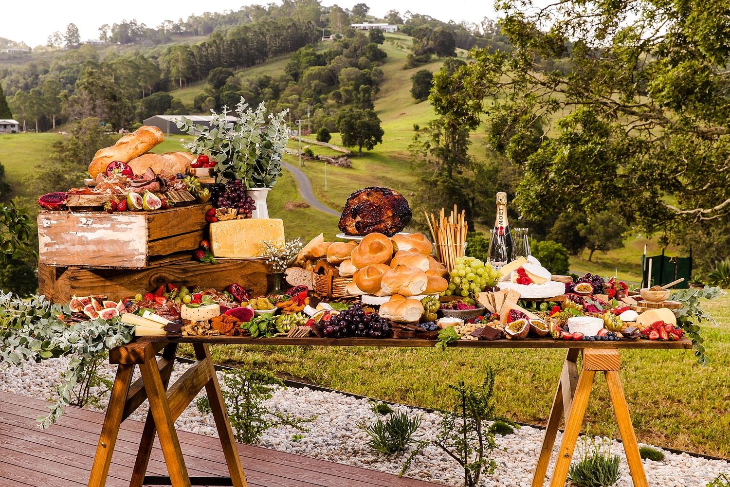 Rustic outdoor wedding reception setup with wooden tables and chairs on a grassy hillside at sunset.