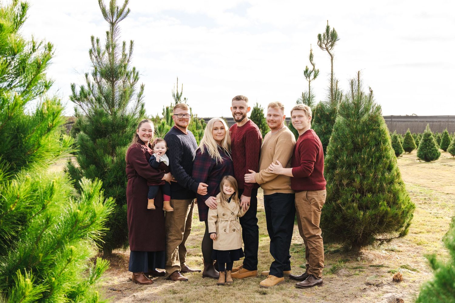 Family posing together at a Christmas tree farm surrounded by lush green pine trees on a sunny day.