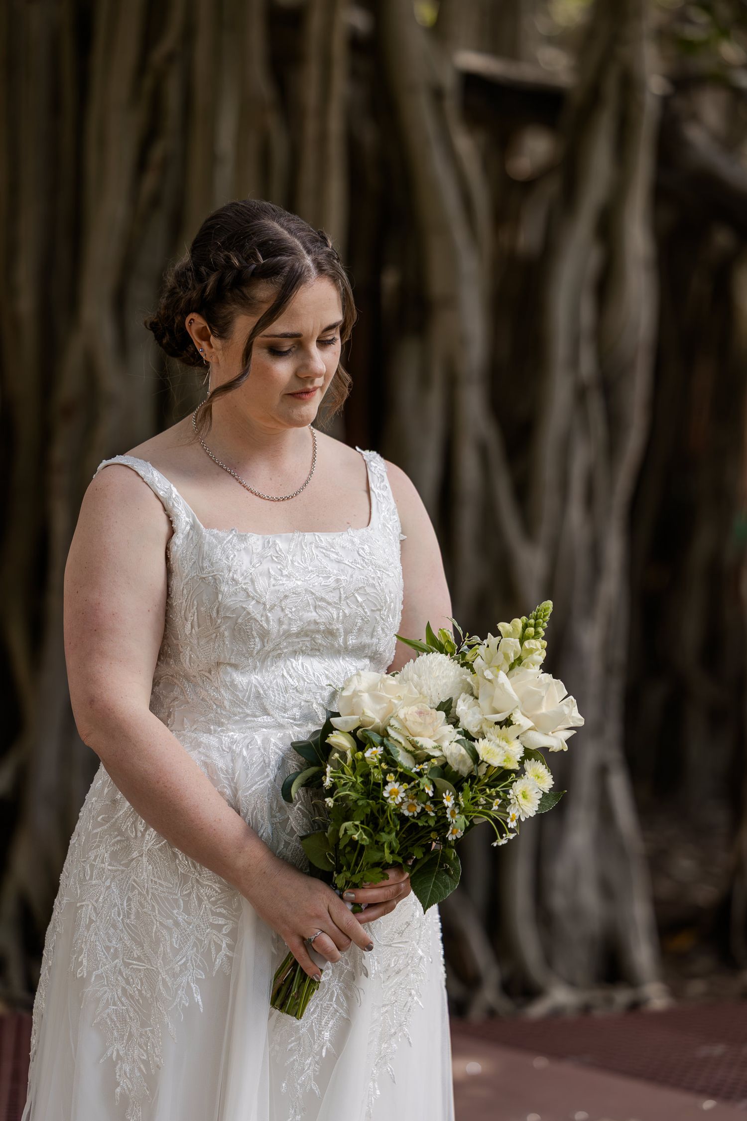 Bride in white lace gown holding white floral bouquet, standing before a large banyan tree backdrop.
