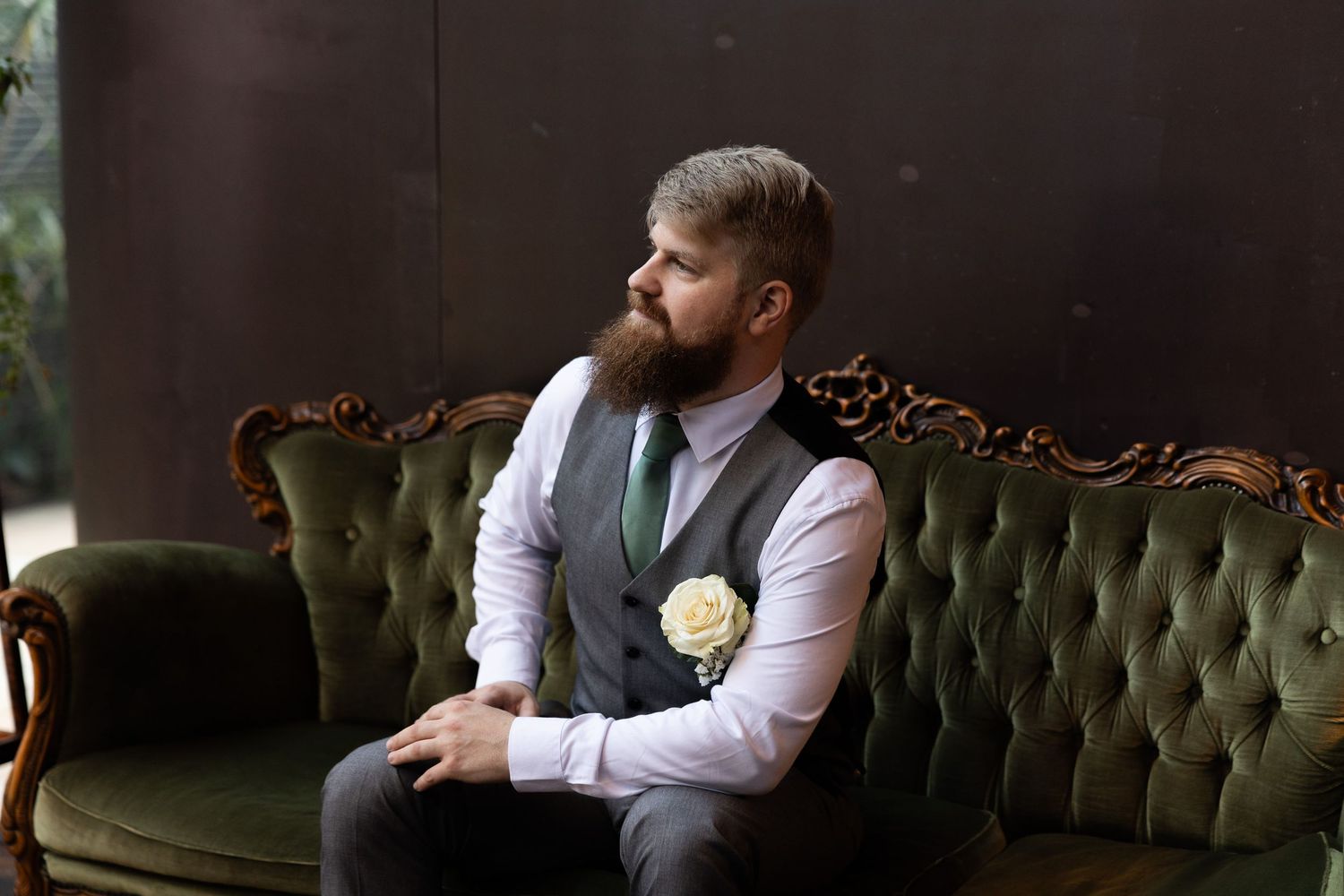Bearded groom in gray vest and white shirt with boutonniere sitting on green velvet sofa.