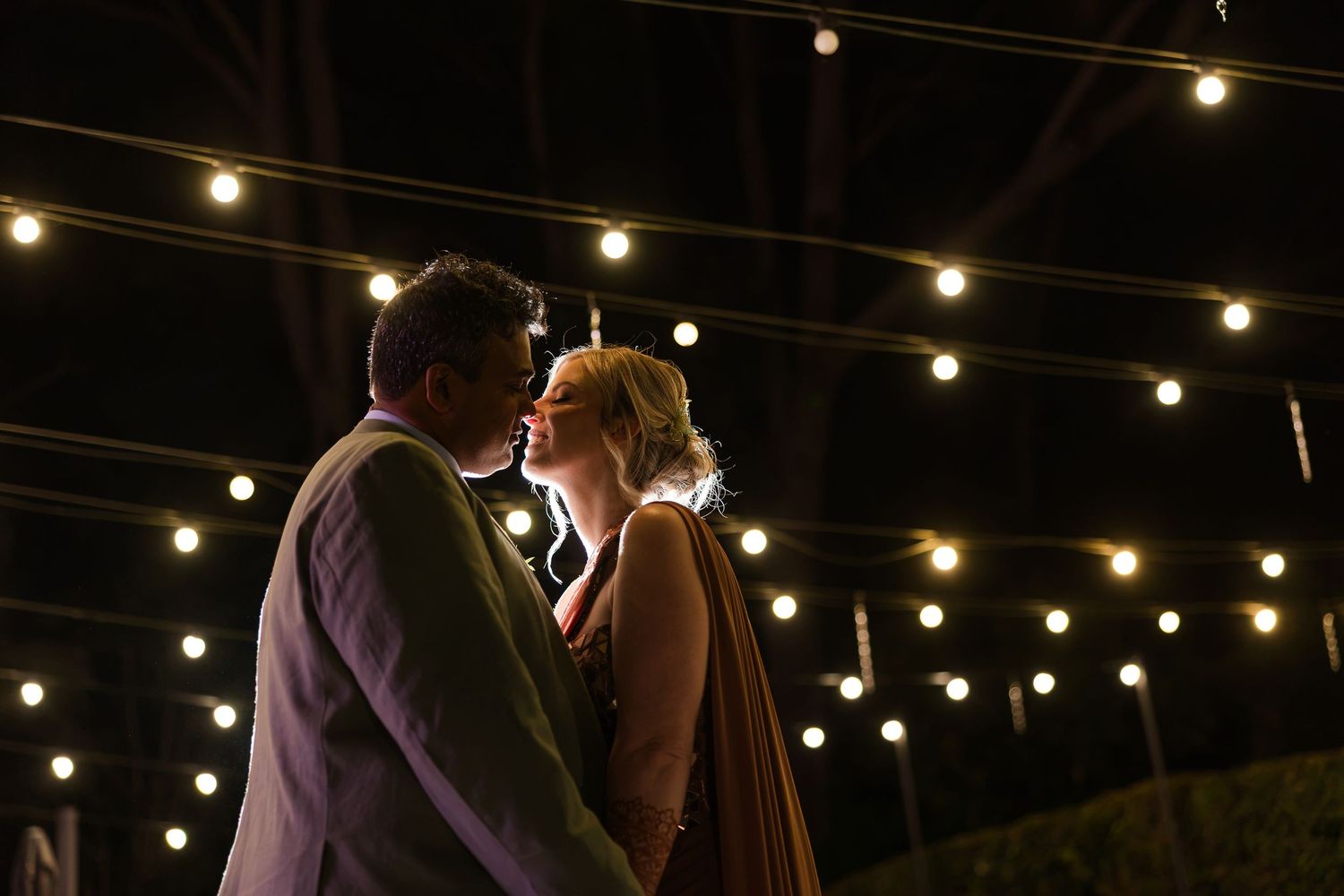 Couple sharing a romantic moment under glowing string lights at an outdoor evening event.