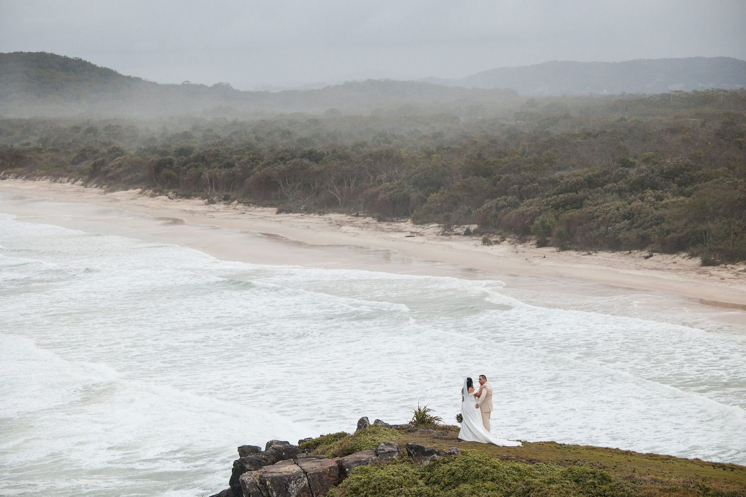 Bride and groom standing on rocky coastal cliff overlooking a misty beach with crashing waves and lush greenery.