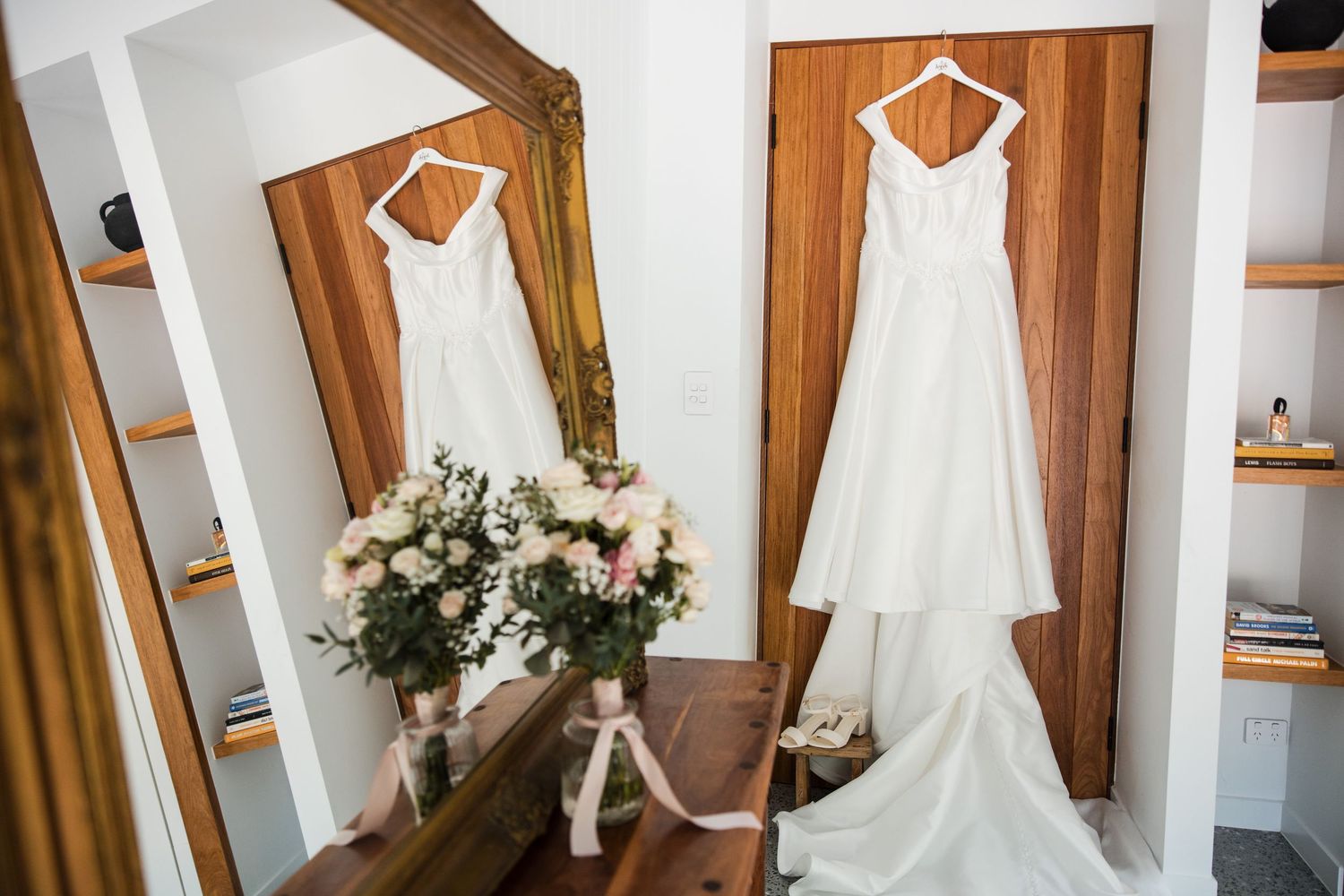 White wedding dress hanging near a gold mirror with bridal bouquets of pink and white flowers in an elegant bridal suite.