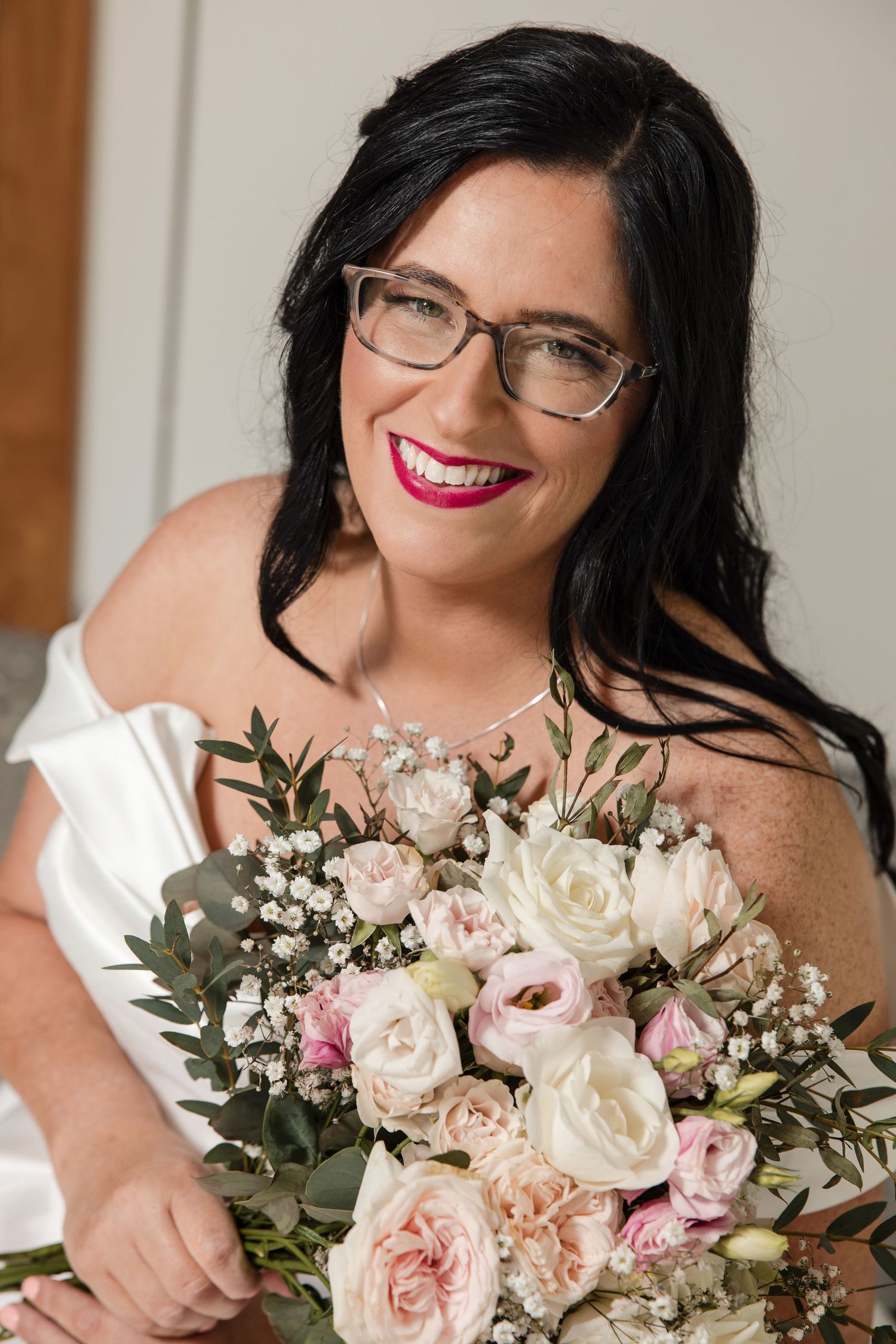 Smiling bride with dark hair and glasses holding a bouquet of pink and white roses in a white off-shoulder dress.