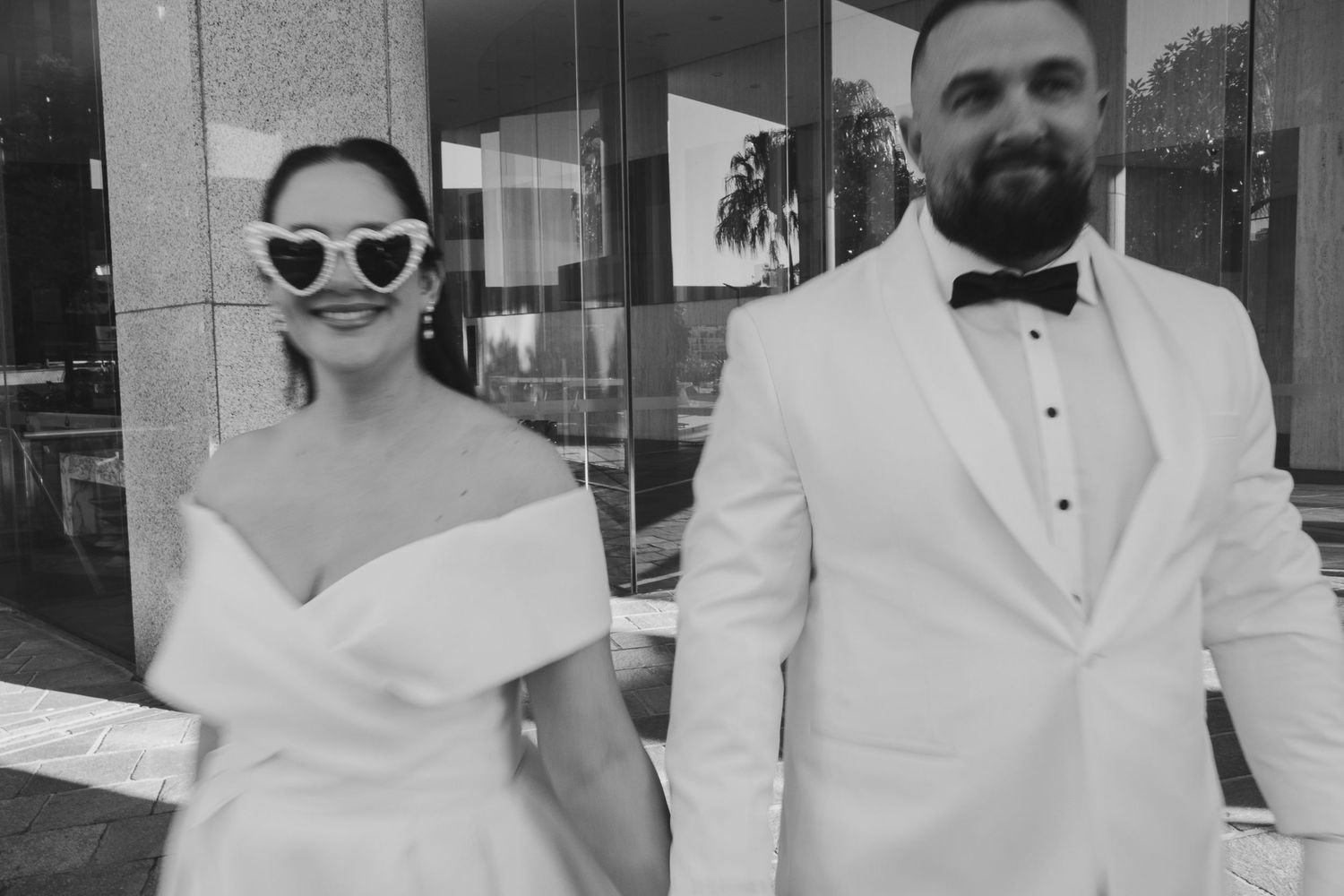 A couple in matching white formal attire walks together, with the bride wearing sunglasses and an off-shoulder gown.