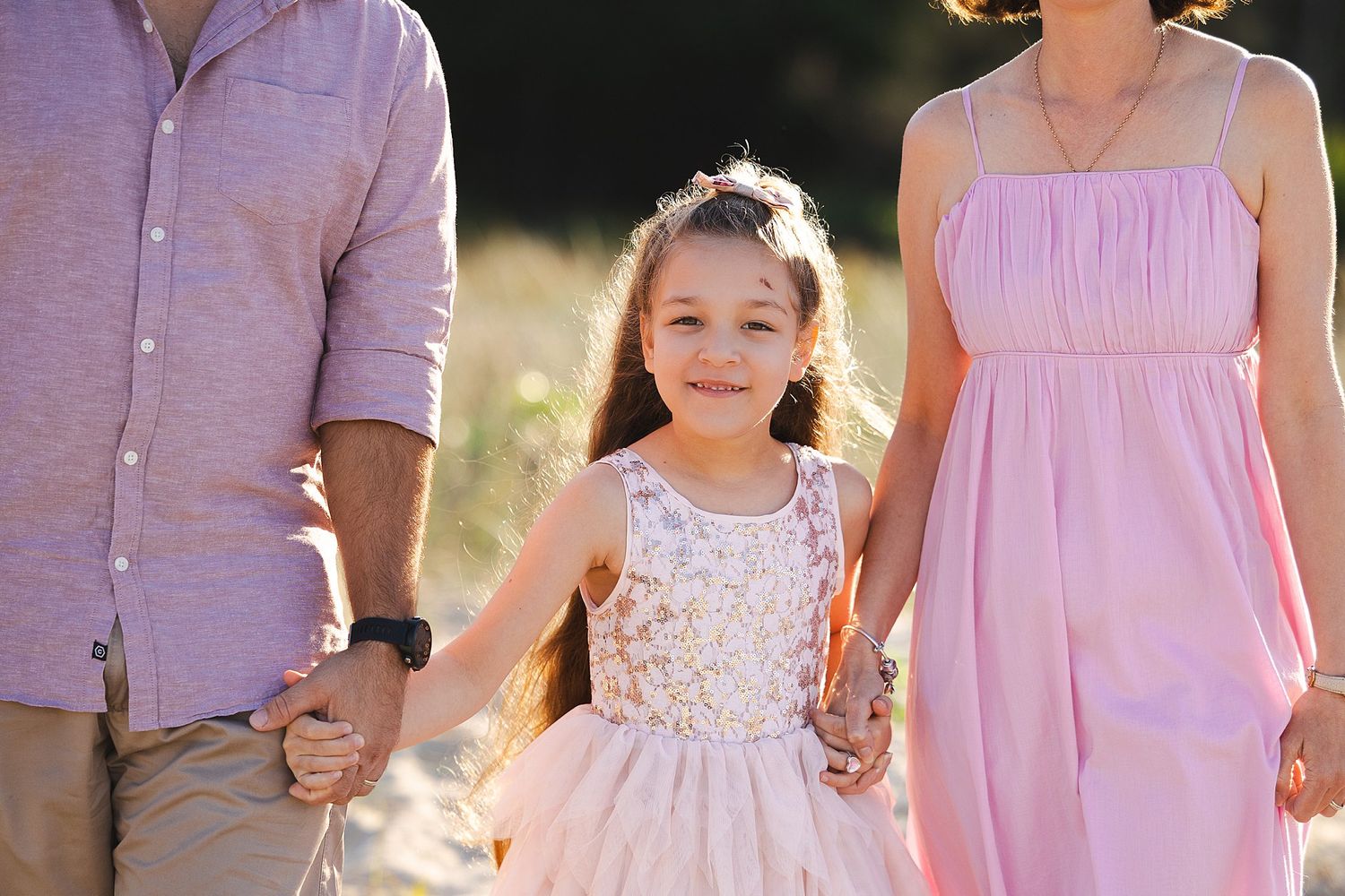 Three people stand together in a sunny field wearing pink and white clothing for a family portrait.
