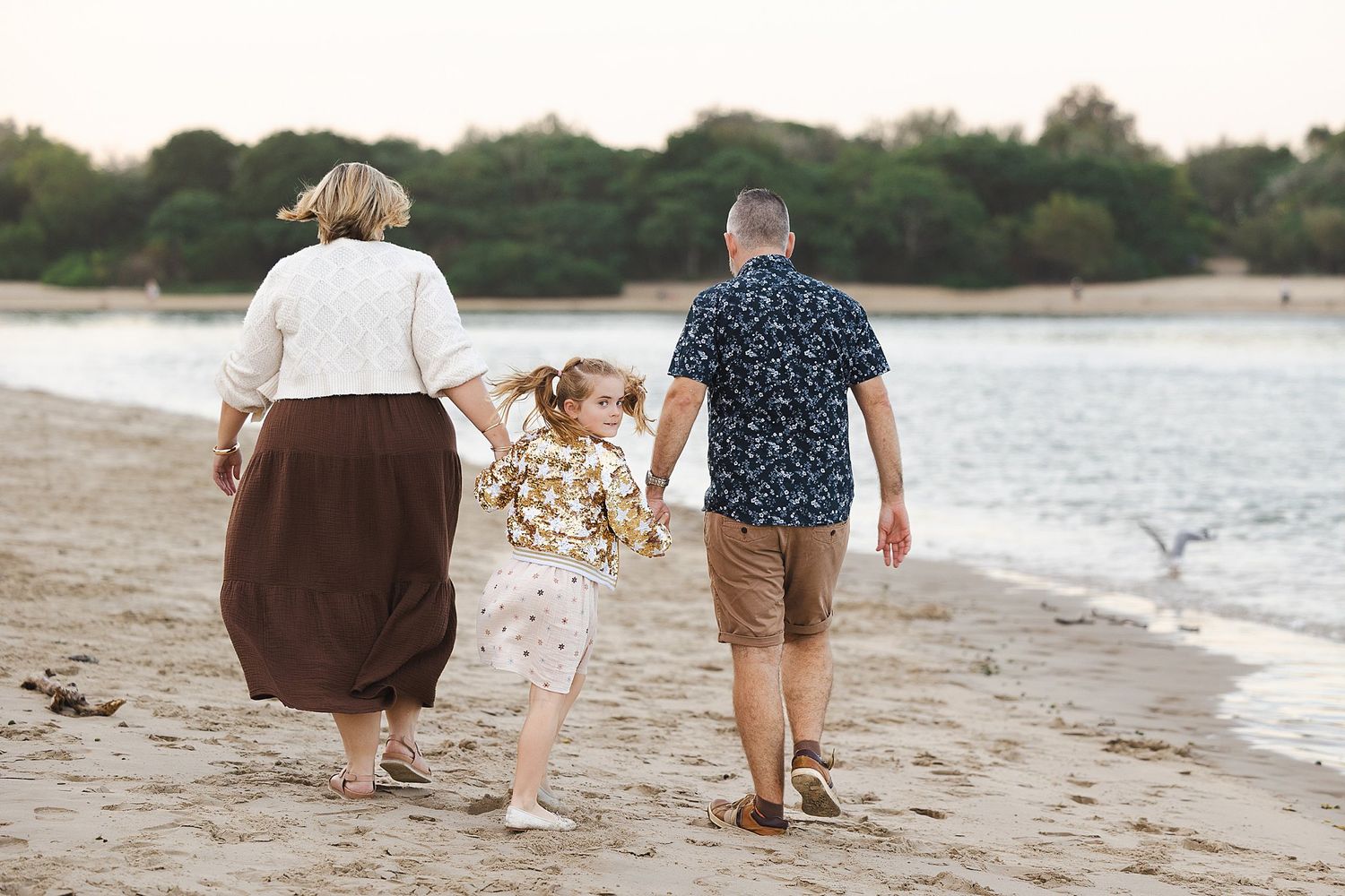 Three people walking and holding hands together along a sandy beach at sunset with water and trees in background.