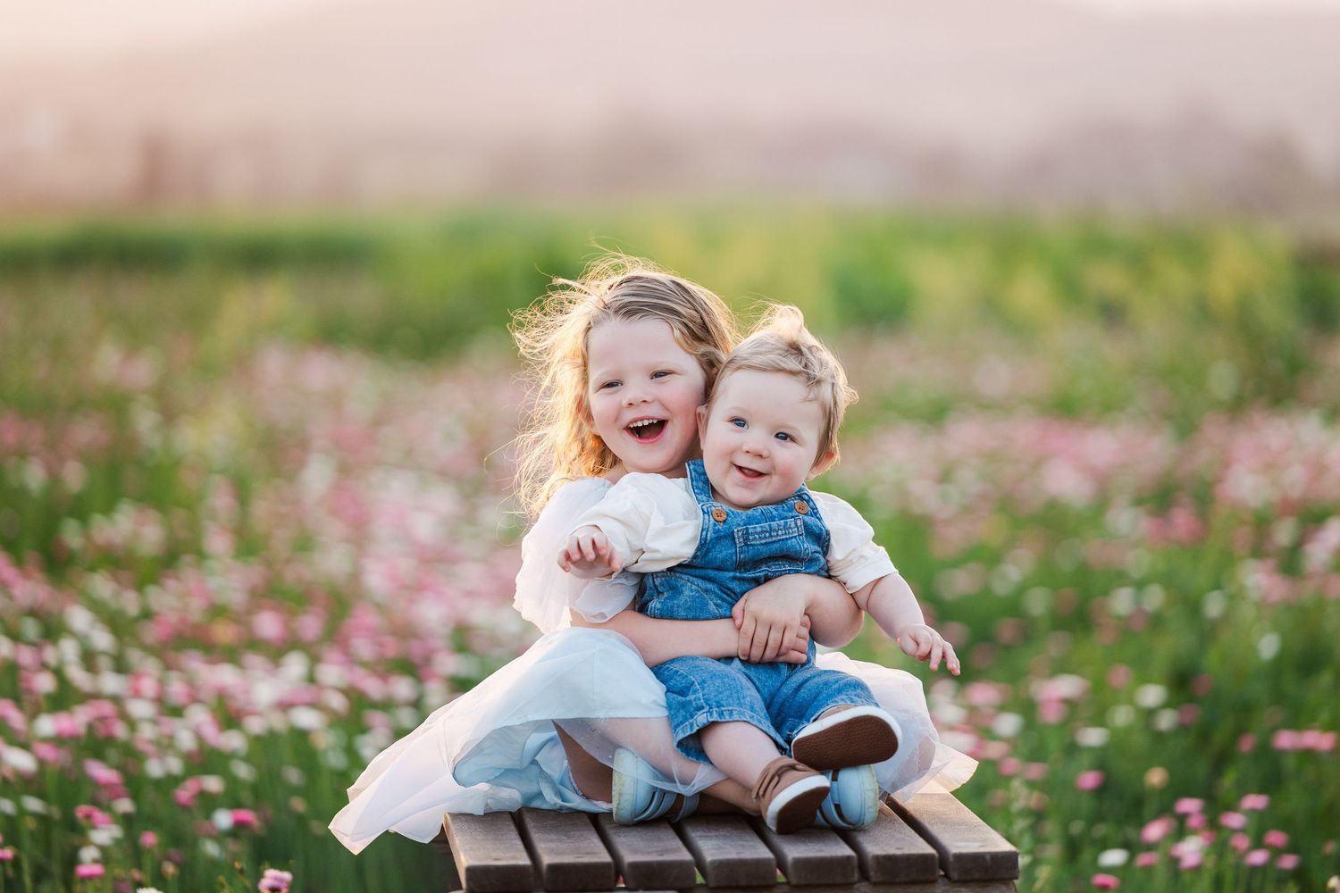 Two children share joyful laughter while sitting together on a wooden bench in a field of pink wildflowers.