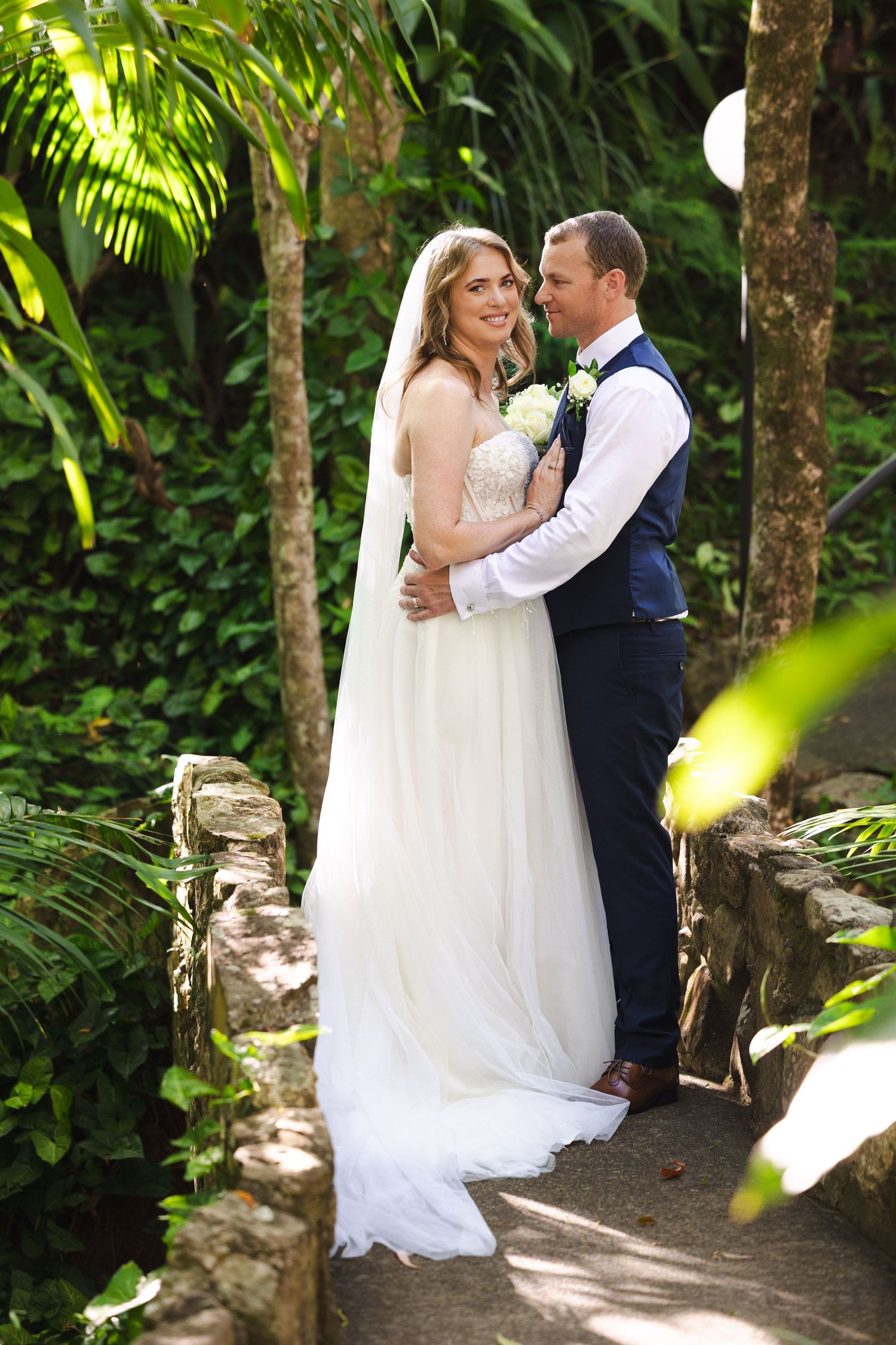 Wedding couple embraces in a sunlit garden pathway surrounded by tropical foliage and trees.