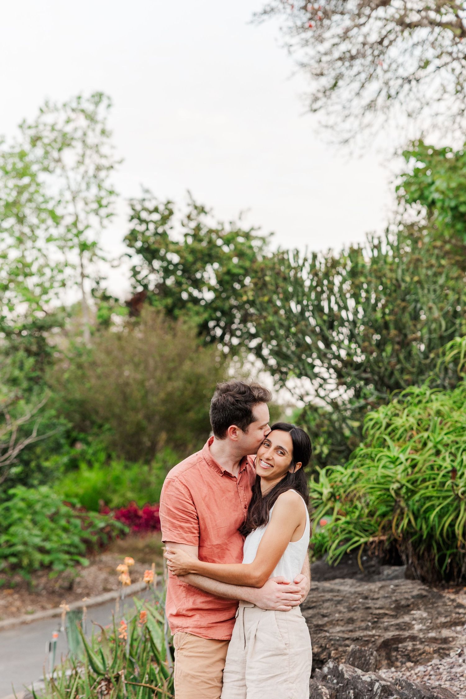 A couple embraces outdoors in a garden setting with blooming plants and greenery in the background.