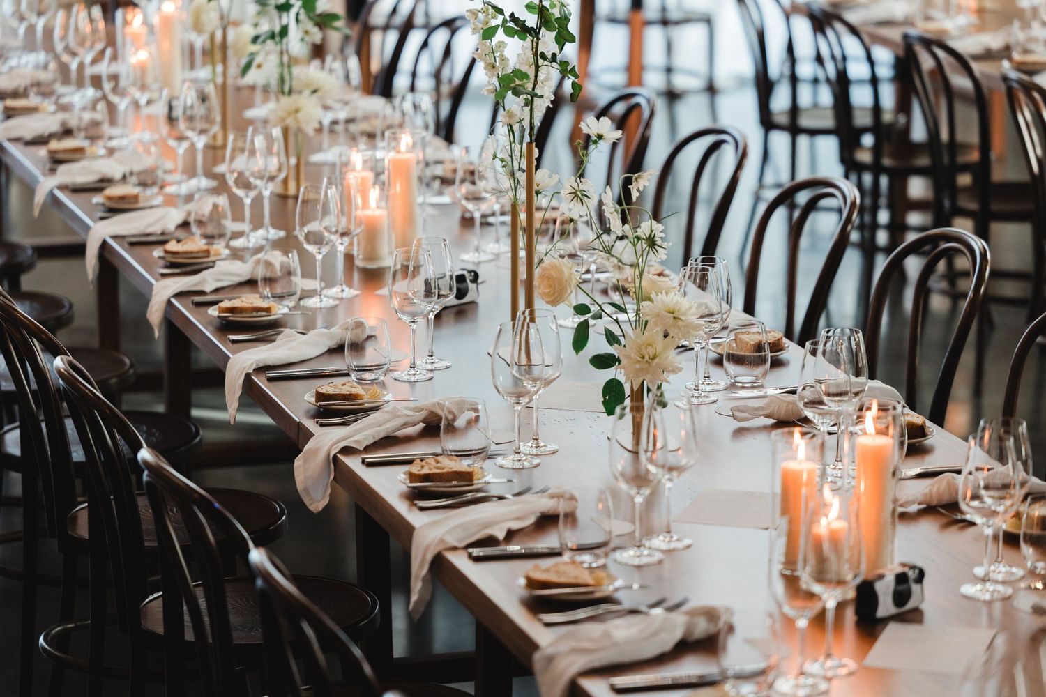 Elegant wedding reception table set with glowing candles, white linens, and bentwood chairs in a rustic setting.