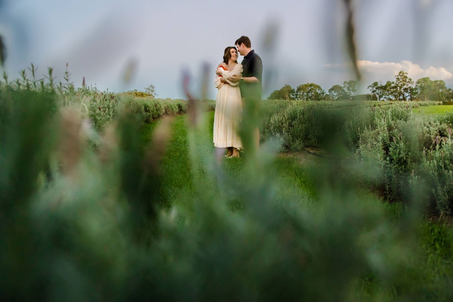 A romantic couple embraces in a lush green field on their wedding day, viewed through soft foreground foliage.