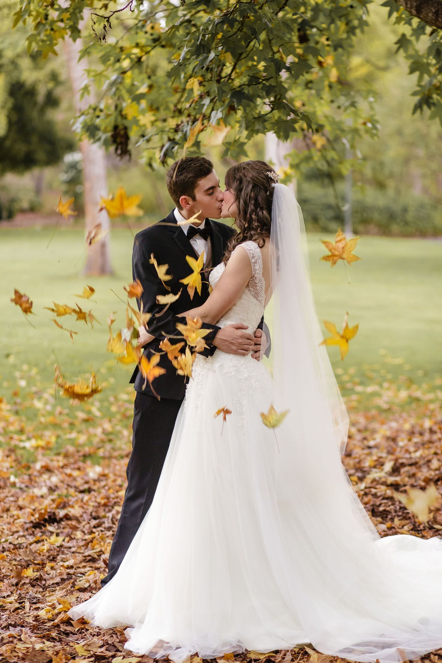 A romantic autumn wedding photo with falling yellow leaves as a couple embraces outdoors in formal attire.