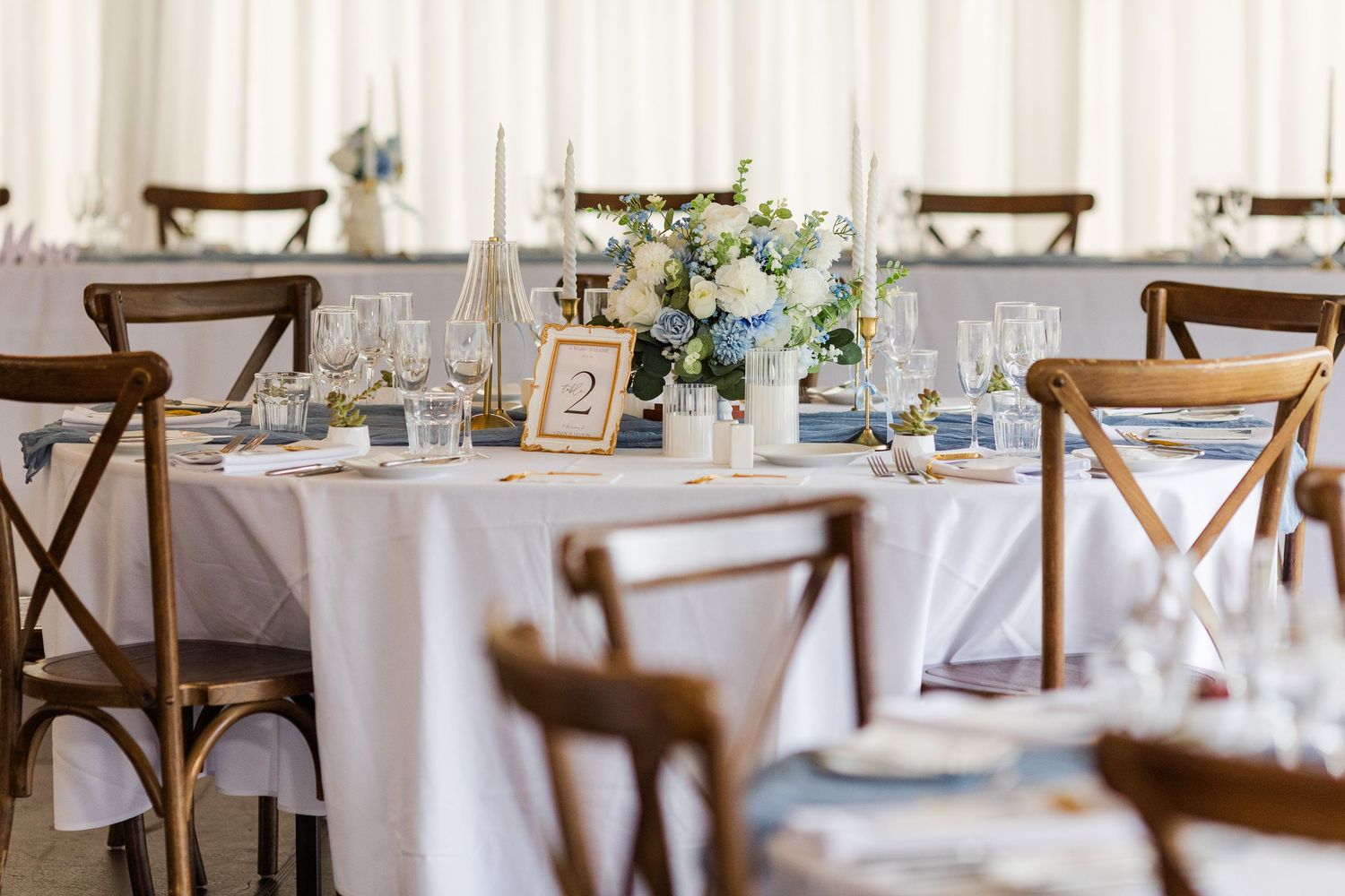 Elegant wedding reception table with white linens, wooden cross-back chairs, and blue and white floral centerpiece.