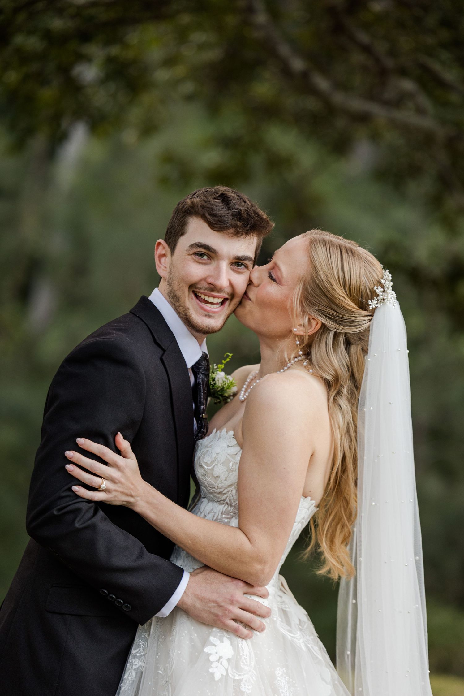 A smiling groom in a black tuxedo receives a kiss on the cheek from his blonde bride in a lace wedding gown.