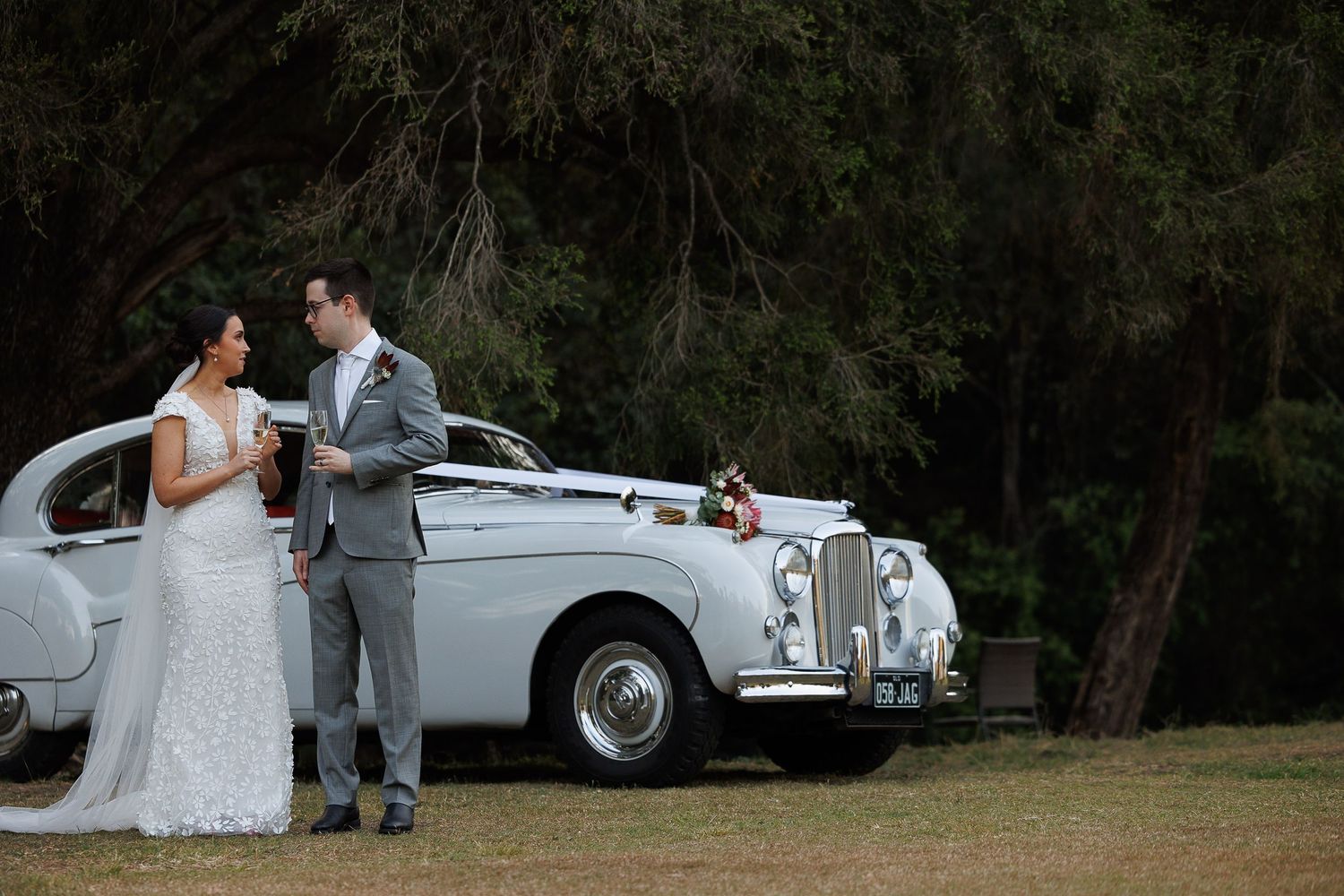 Bride and groom standing beside a classic white vintage car surrounded by lush green trees on their wedding day.