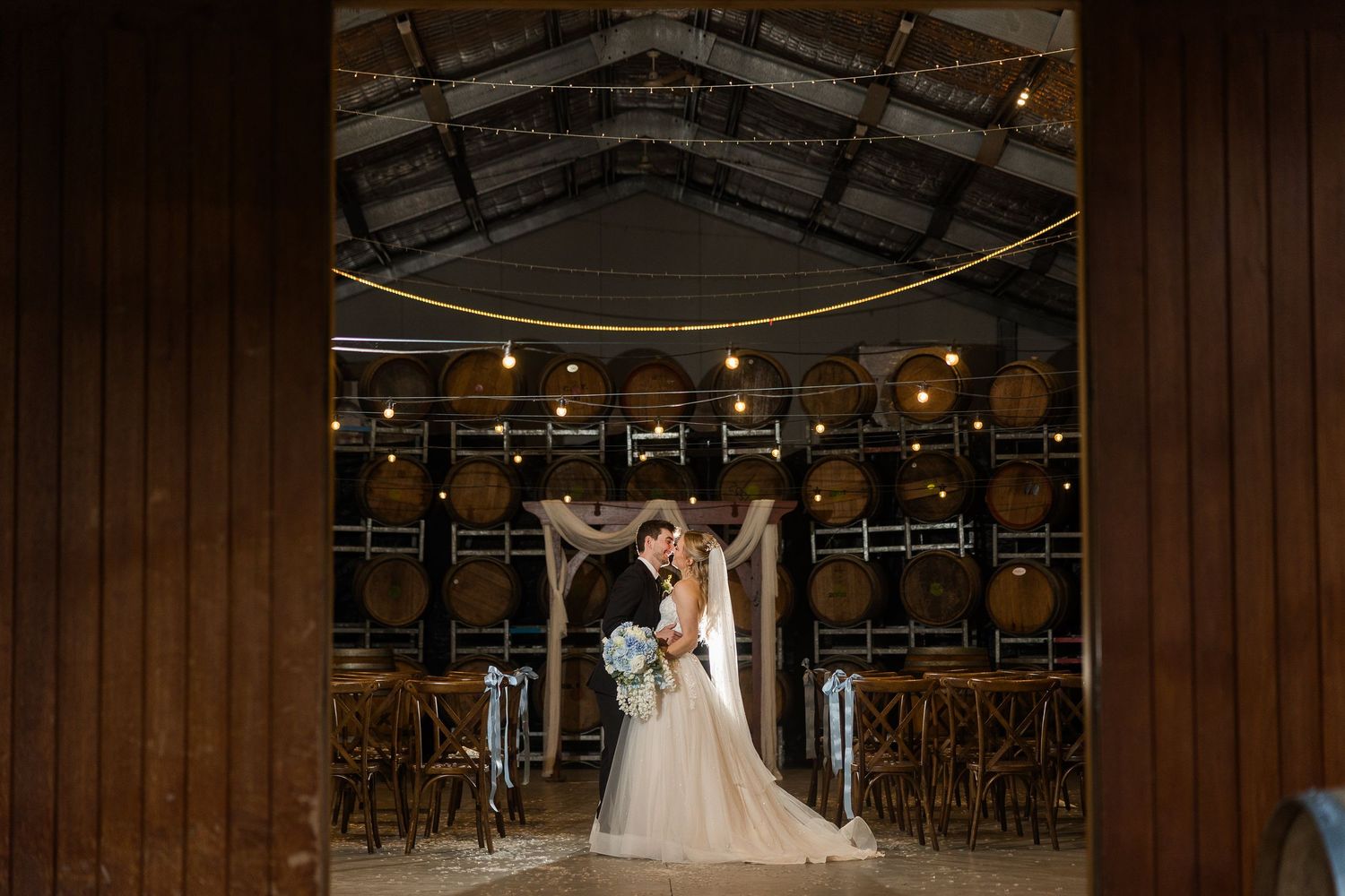 Bride and groom share a romantic kiss in a rustic winery venue with wooden barrels and warm string lights.