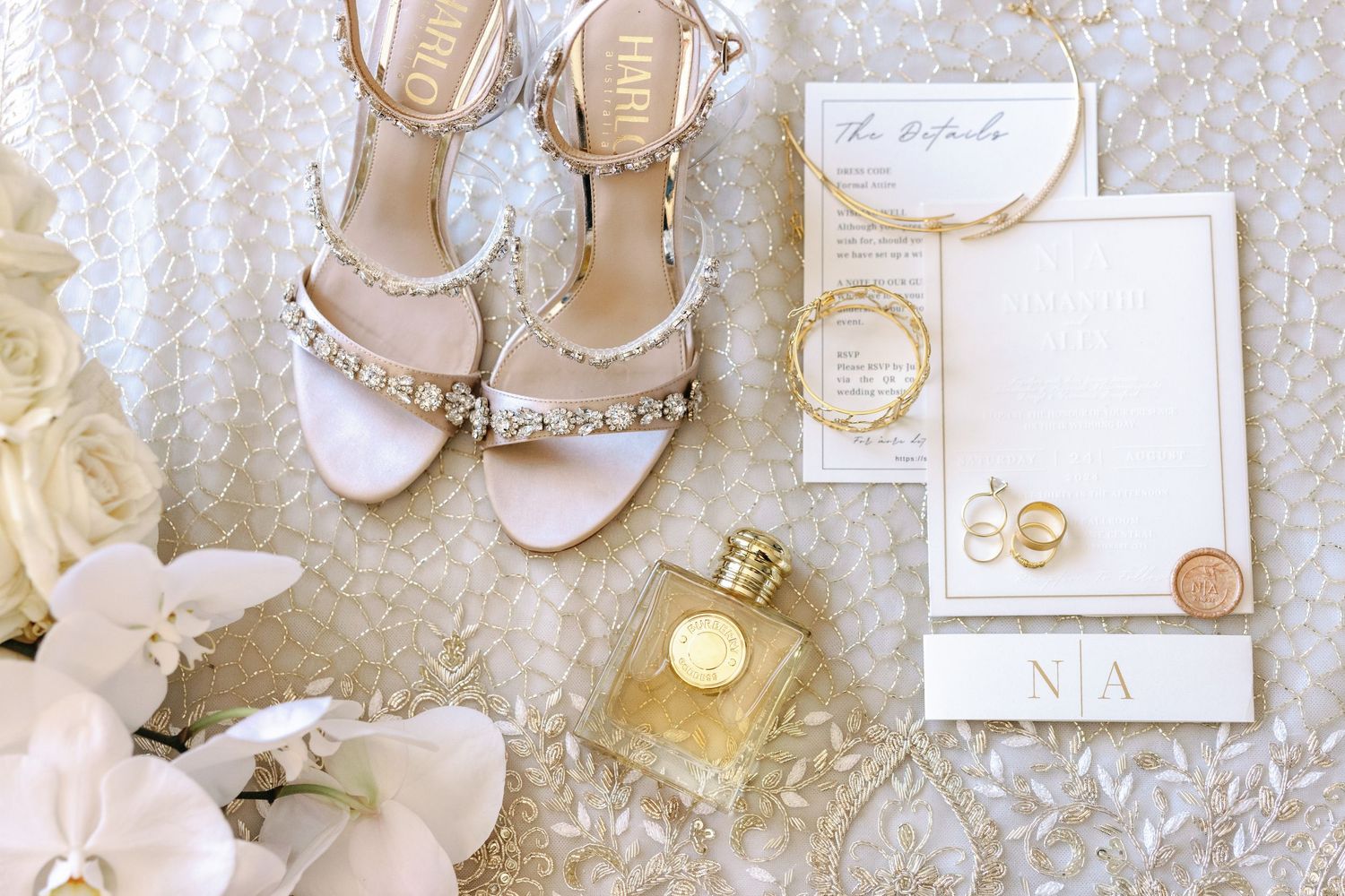 Elegant wedding accessories including white shoes, jewelry, and invitations arranged on lace background.
