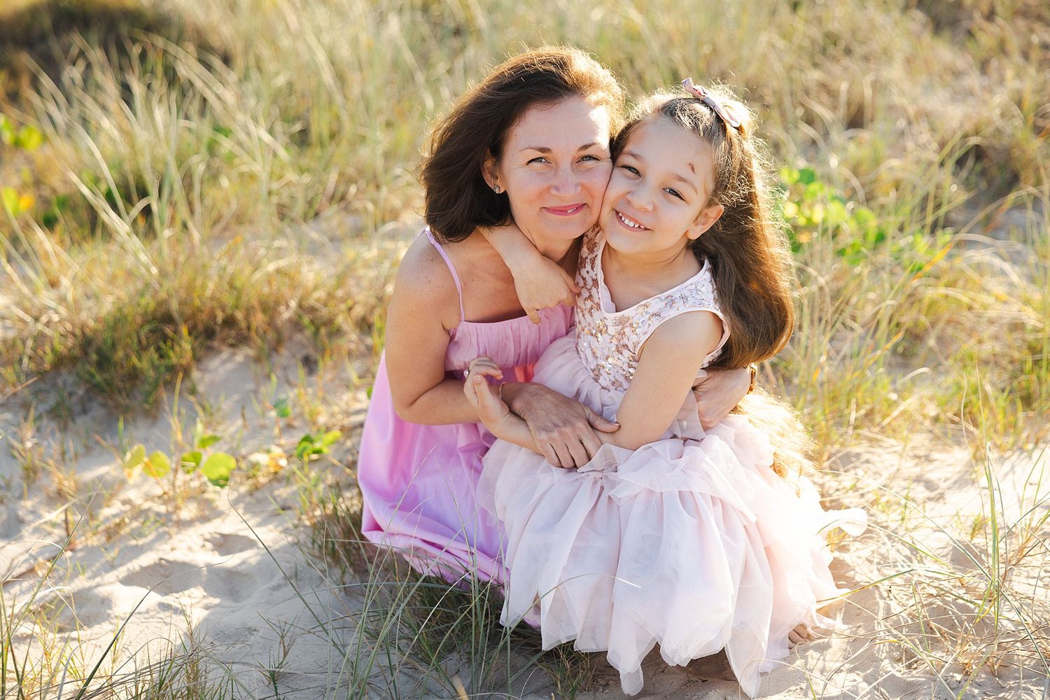 Two people wearing pink dresses embrace while sitting in tall grass during golden hour.