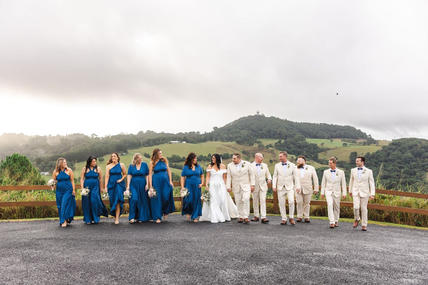 Wedding party walks outdoors with bridesmaids in teal gowns and groomsmen in tan suits against misty green hills.