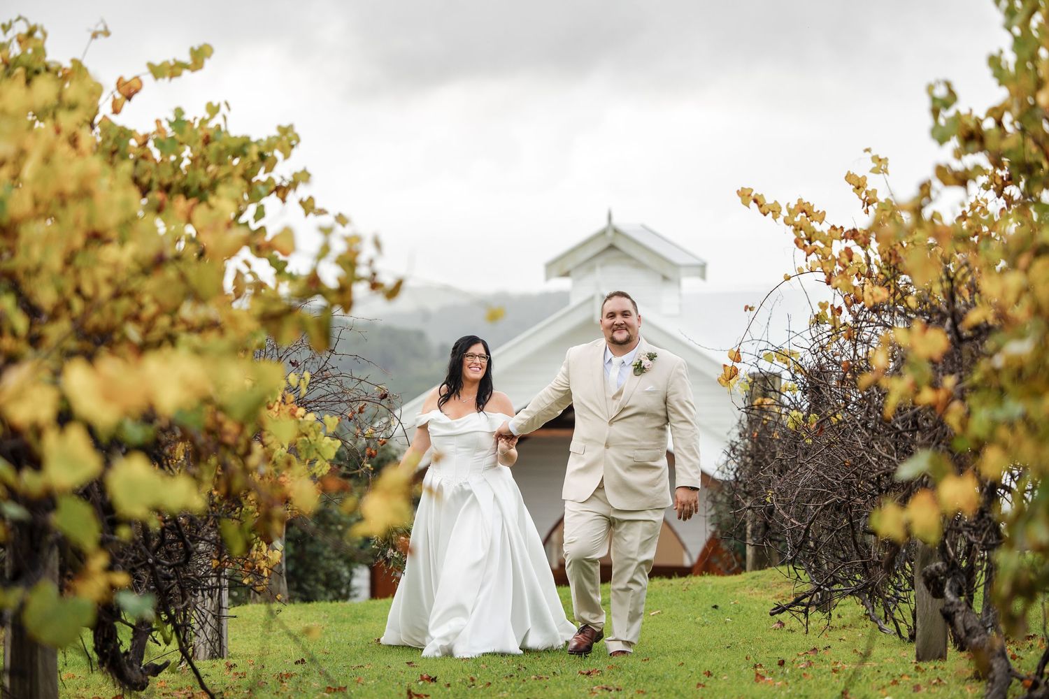 Bride and groom walking hand in hand through a vineyard with a white chapel and rolling hills in the background.