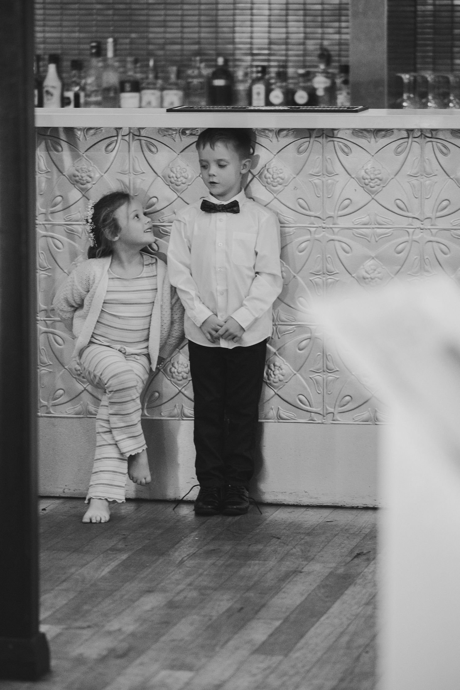 Black and white photo of two dressed-up children, a girl in stripes and a boy in bow tie, standing by a decorative wall.